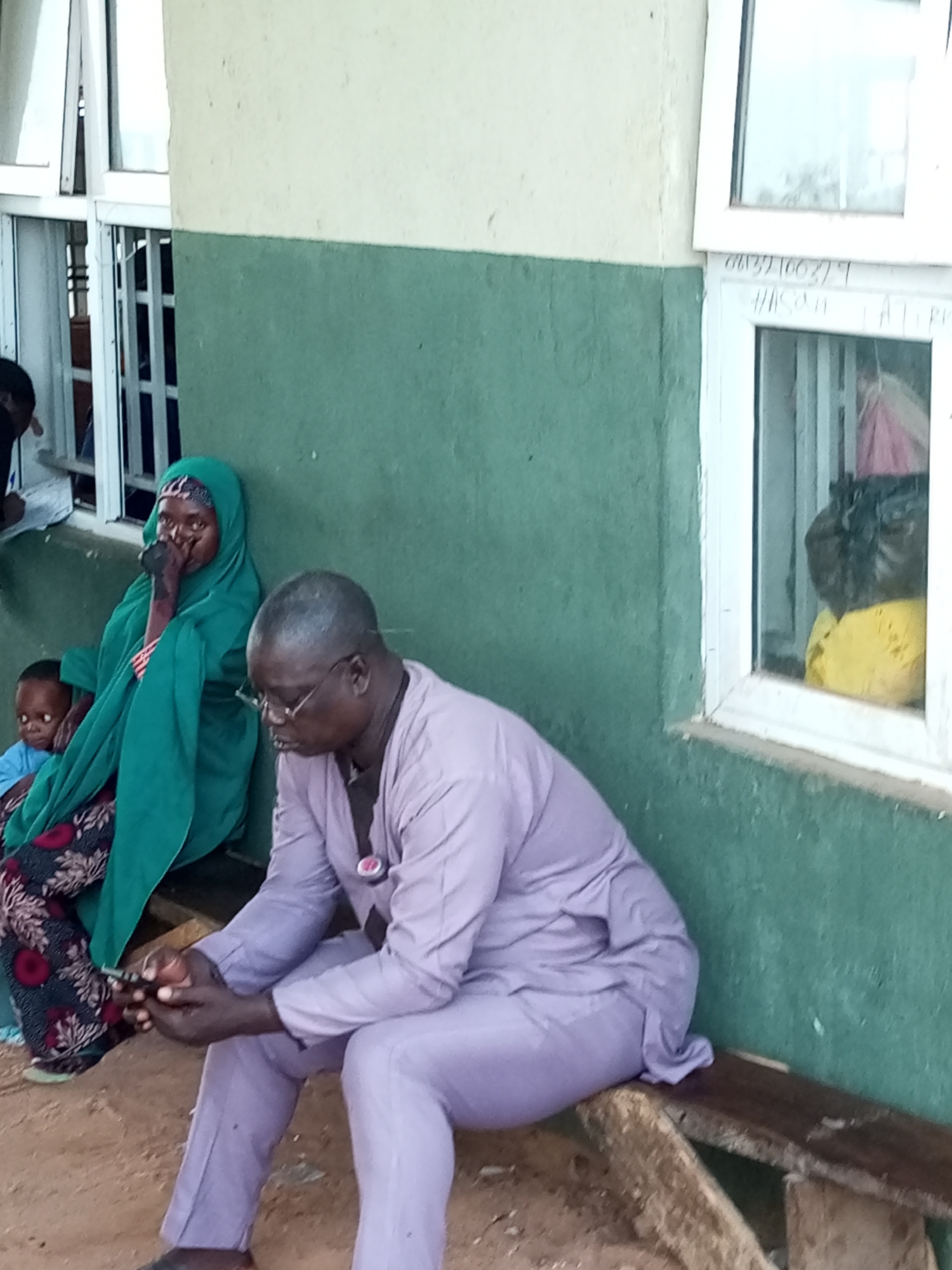 In Gombe, Nigeria, an outdoor scene depicts a group of people near a building with windows. In the foreground, a man wearing glasses and a light purple traditional outfit sits on a wooden bench, holding a mobile phone. To his left, a woman in a green hijab and patterned dress is seated, resting her head on her hand and appearing to be speaking or in distress. A young child in a light blue shirt is seated in front of her, looking towards the camera. Behind the seated individuals, a building wall is visible, painted in two tones: a lighter color on the top and a darker green on the bottom. Two windows are present, one showing reflections and what appear to be items inside, including yellow and dark-colored bags. The ground is sandy and uneven. The overall impression is a moment of waiting or reflection in a public or semi-public space.