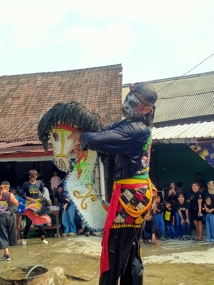 The image captures an outdoor cultural performance in Sepat, Indonesia, occurring on wet, muddy ground under a clear sky. A central performer faces left, clad in a wet black long-sleeved shirt, dark pants, a multi-colored checkered sarong, and a draped red and yellow sash. Their face is painted with white, black, and red patterns resembling a skull or monster, featuring prominent ears and a dark fabric head covering with a braided band. The performer holds a large, flat, decorative white horse effigy with a black shaggy mane, adorned with red, green, and gold patterns. To the left, another individual in a black shirt with a graphic print holds a smaller, red and black feathered object near a wooden table. In the background, a crowd of spectators, including adults and children mostly in black attire, observes the event, with some utilizing mobile phones for photography. The environment includes structures with red tiled and corrugated metal roofs in the background. A rusted metal barrel with a hose is visible in the lower-left foreground.