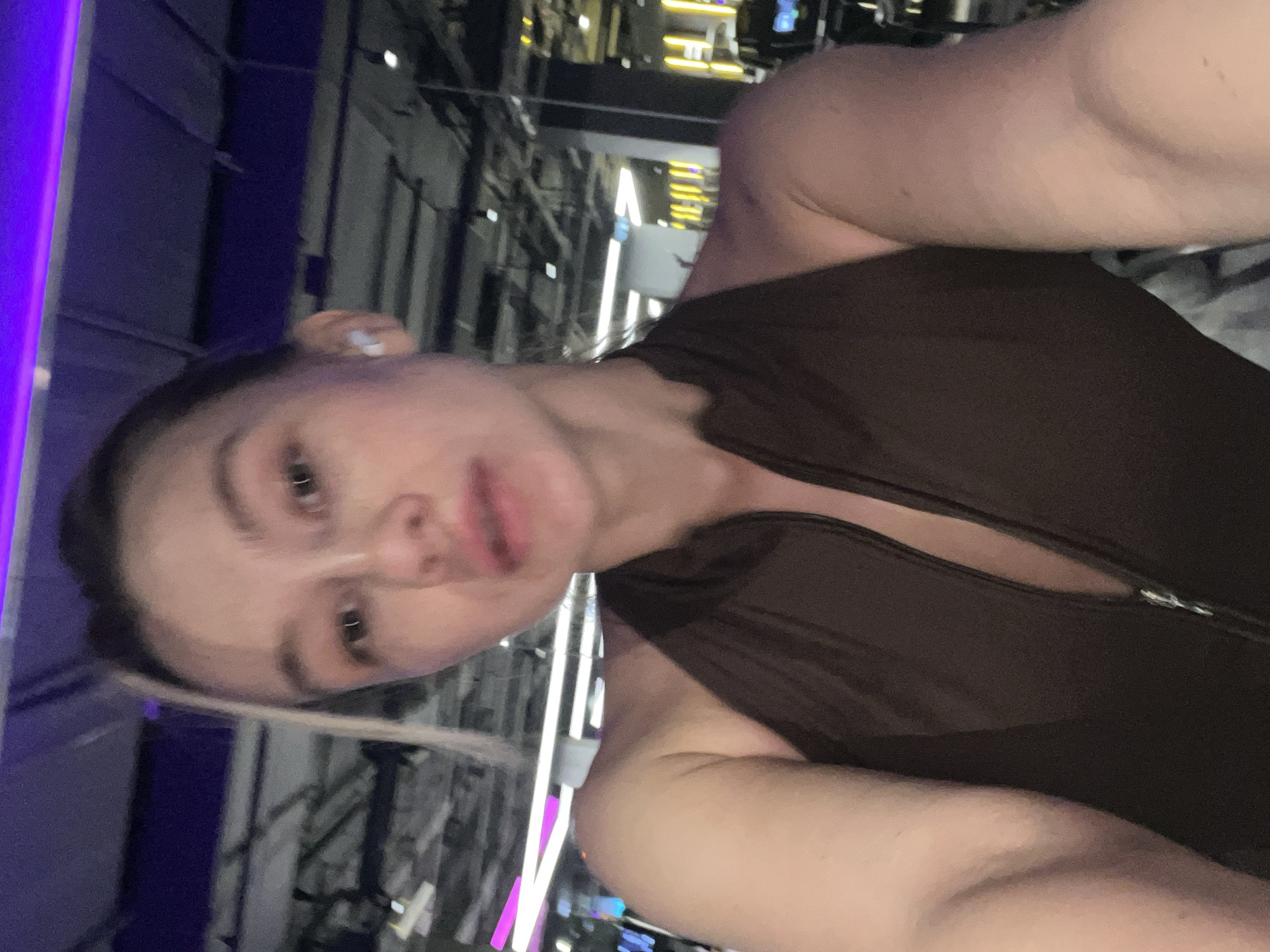 A selfie photograph shows a person in an indoor setting with linear architectural elements in the background. The person is wearing a dark brown garment with a zipper. An earbud is visible in the person's left ear. The location is identified as Voronezh, Russia.