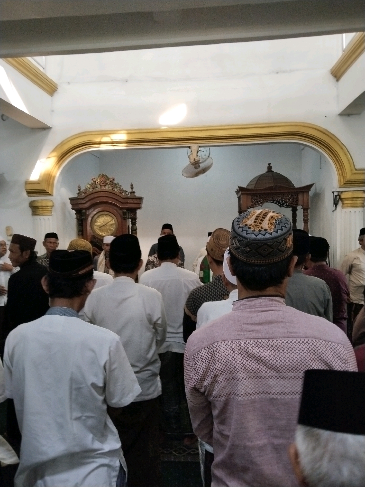 A group of men, mostly wearing peci hats and white shirts or kurtas, are gathered inside a building with ornate gold trim along the ceiling arches. The men appear to be in prayer or a religious service, with their backs to the camera, facing a decorative wooden structure with a domed top and a large grandfather clock with a round face. Sunlight streams in from above, illuminating the room. The scene likely takes place in a mosque or religious hall in the city of Serang, Indonesia, given the attire and religious context.