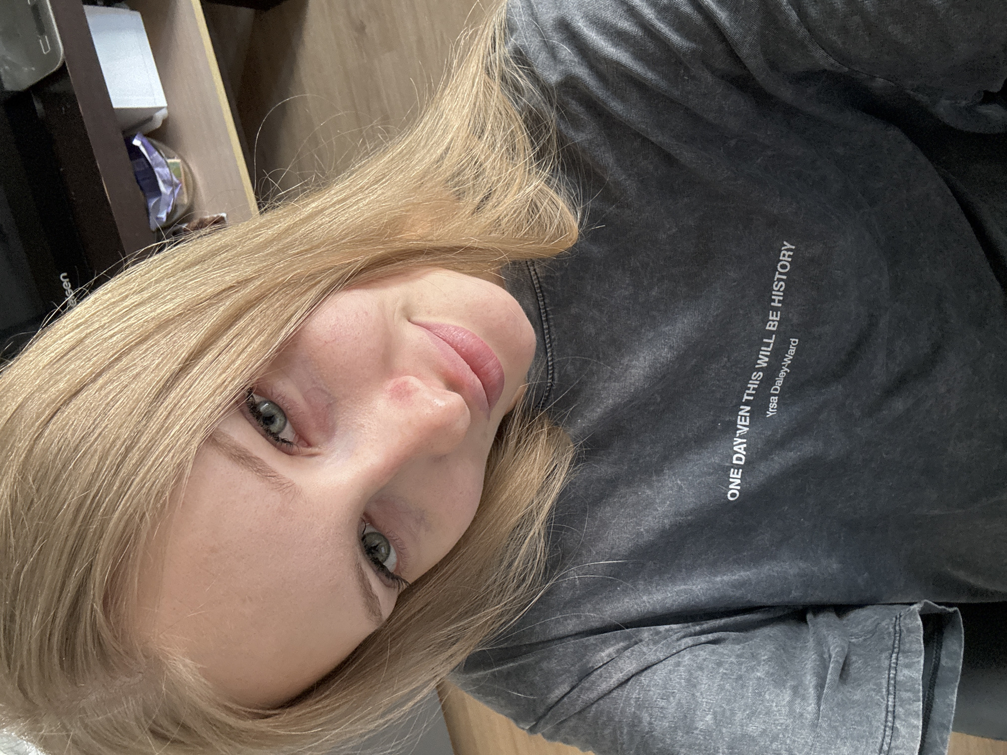A young woman with blonde hair and green eyes is taking a selfie. She is wearing a dark grey t-shirt with white text that reads "ONE DAY EVEN THIS WILL BE HISTORY" and "Yrsa Daley-Ward". She is looking at the camera with a slight smile. The background is blurred but appears to be indoors, with shelves and some objects visible. There is no indication of the location being in the city of Perm, Russia.