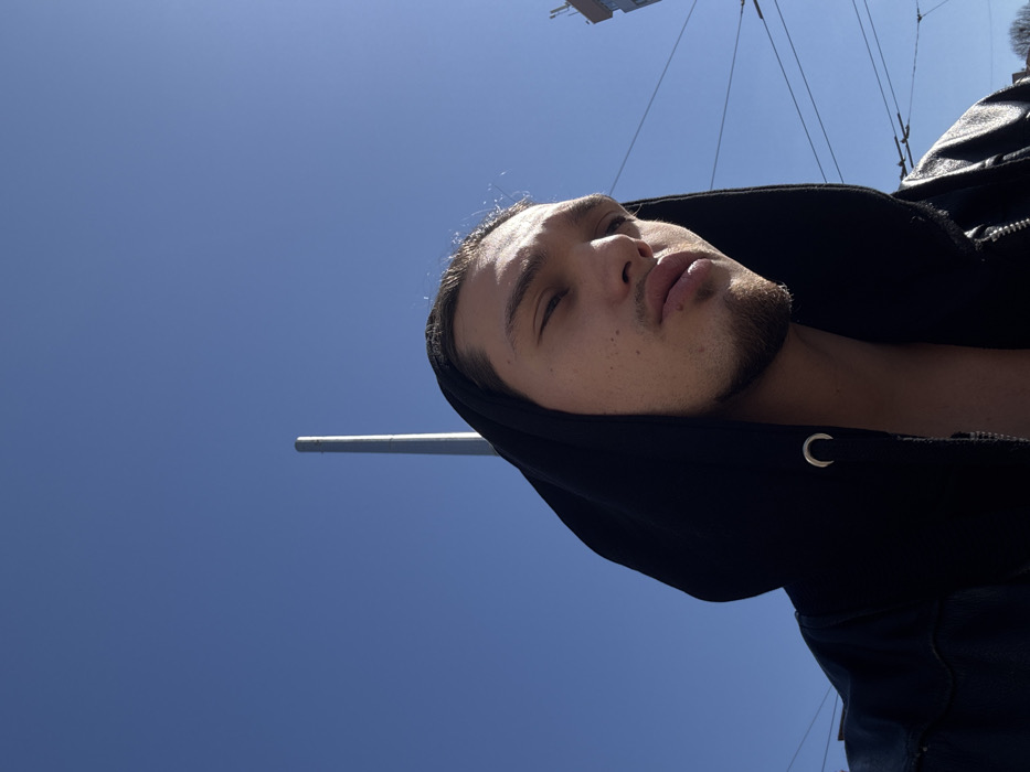 A young adult male is positioned in a close-up, low-angle photograph against a clear blue sky, indicating a bright, sunny day in an urban environment consistent with Sofia, Bulgaria. The male occupies the lower portion of the frame, wearing a black hooded sweatshirt with the hood pulled up and what appears to be a black leather jacket. The sweatshirt is partially unzipped, exposing his chest. He has dark hair and a short beard. His head is tilted slightly upwards and to the right, with his eyes partially squinted, suggesting direct sunlight exposure. His lips are pursed. A tall, light grey vertical pole is visible in the background, slightly left of center, extending towards the top of the frame. Multiple dark overhead wires, characteristic of electric public transport infrastructure, cross horizontally in the mid-right background. A section of a light-colored building is partially visible in the upper-right corner, behind the wires.