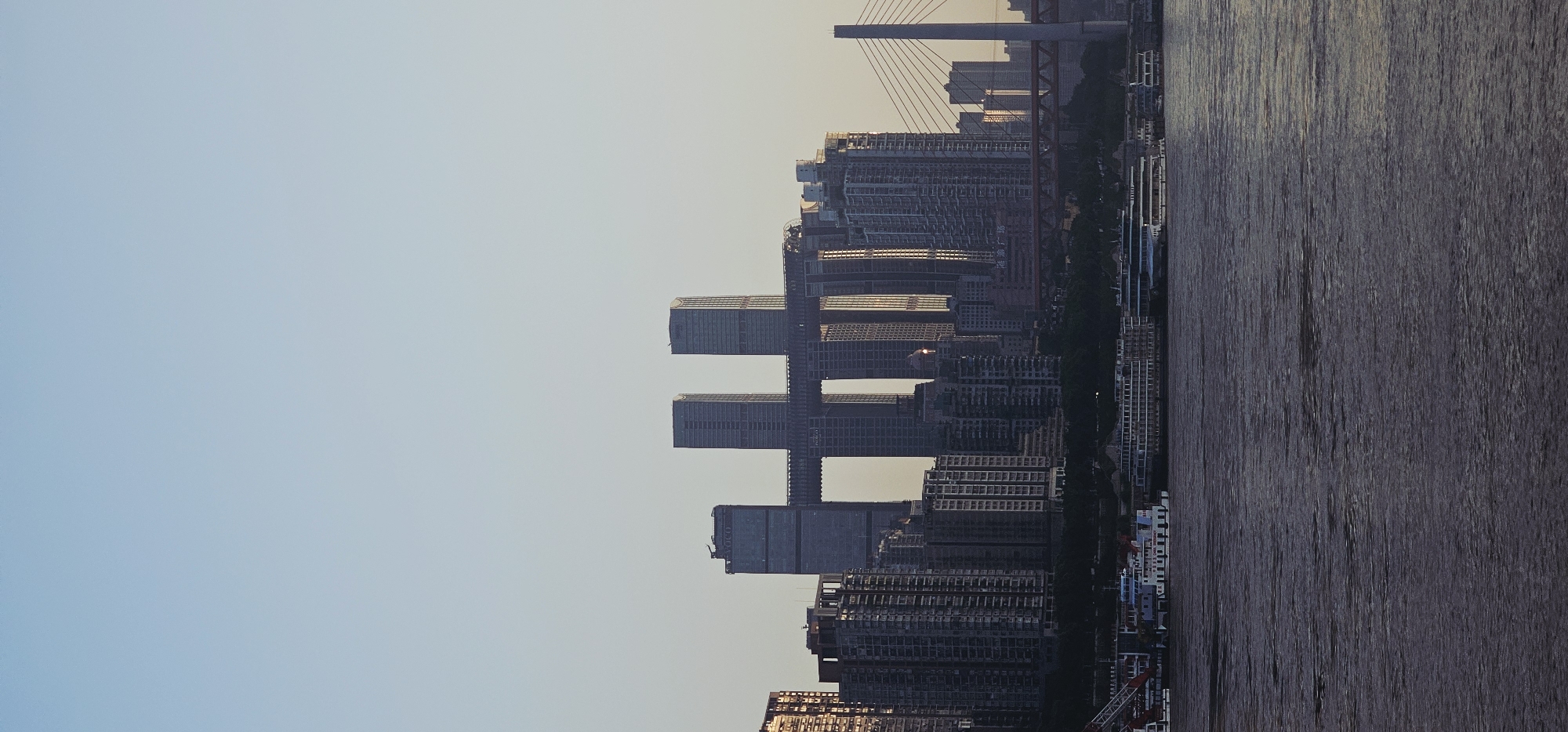 A vertical, medium shot captures a cityscape at dusk along a wide river, likely the Yangtze River in Nan'an District, Chongqing, China. The sky transitions from a pale blue at the top to a lighter hue closer to the horizon. Dominating the scene are several modern skyscrapers, including a prominent structure with a horizontal bridge-like connection between two towers, resembling the "Raffles City Chongqing" complex. Other tall buildings, varying in height and architectural style, line the waterfront.
In the foreground, the river occupies the lower portion of the frame. The water's surface is textured with ripples and reflections, appearing dark and somewhat choppy. Several boats and ferries are docked or cruising along the river. One prominent white ferry with multiple decks is positioned on the left, displaying text that includes "CHINA" and "CHONGQING." Other vessels, including a larger white ship with a long promenade deck, are visible further to the right.
The lighting suggests either sunrise or sunset, with a warm glow illuminating some of the buildings, casting long shadows and highlighting the architectural details. The overall impression is one of urban development and a bustling waterfront. No people are discernible in the image.