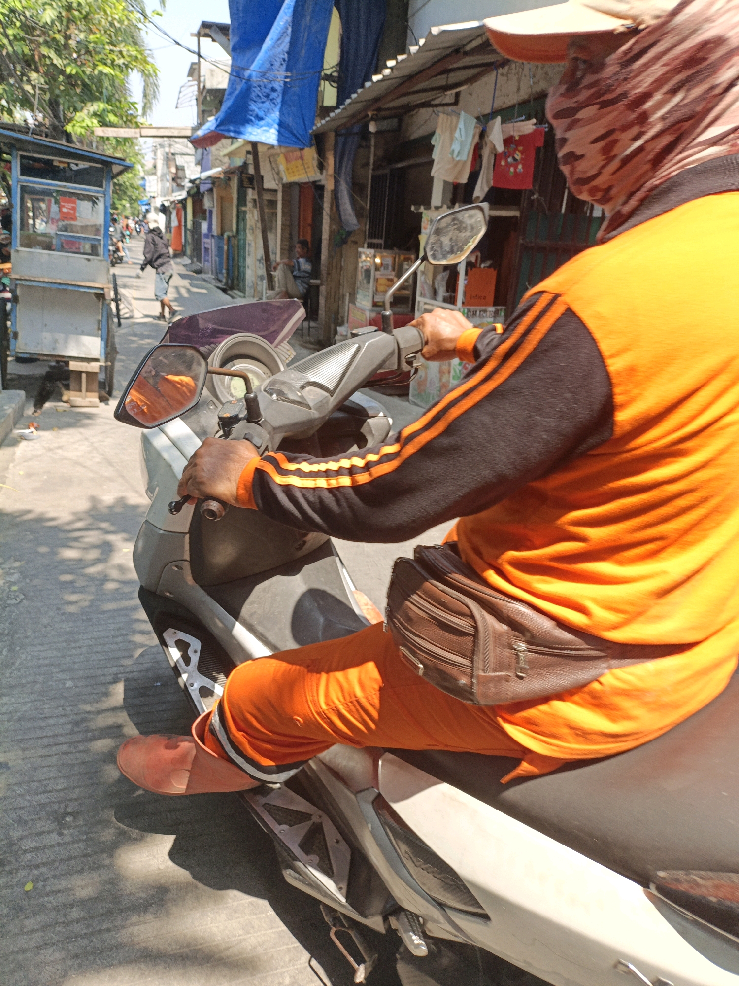 The image shows a person riding a scooter on a street in West Jakarta, Indonesia. The person is wearing an orange long-sleeved shirt with black and orange stripes on the sleeves, orange pants, a brown fanny pack, and a face mask that covers their head and face with a camouflage pattern. The scooter is gray and white. The street is paved with concrete and has shadows from the sun. In the background, there are several small shops and stalls, some with blue tarps over them. There are also trees and other buildings visible. Other people and scooters can be seen in the distance. The scene appears to be a busy urban environment.