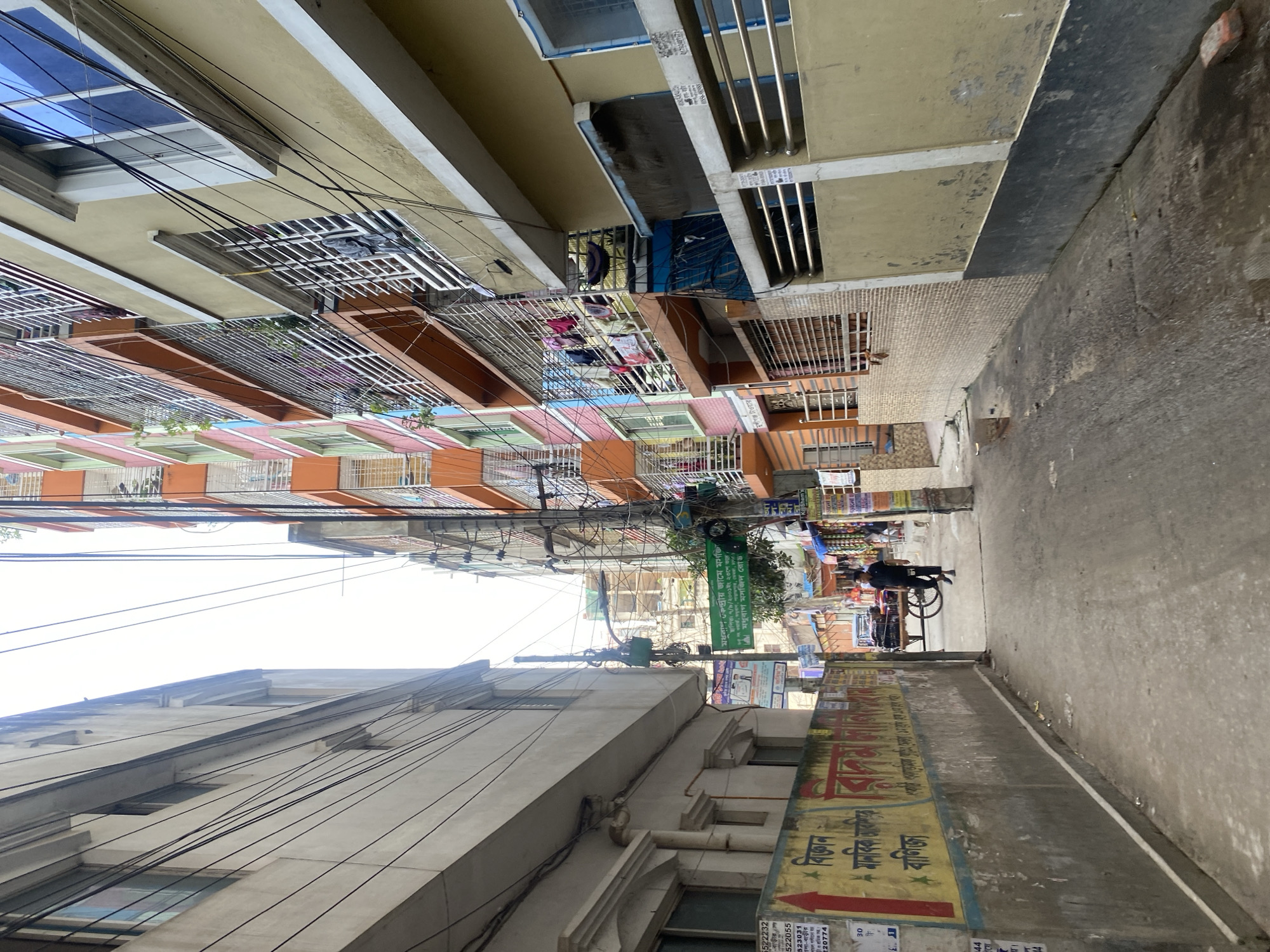 The image captures a street scene in Dhaka, Bangladesh. It showcases the typical urban environment with dense housing. Buildings of various colors and sizes line both sides of the narrow street. Numerous electrical wires crisscross overhead. On the right side of the street, there is a weathered wall with Bengali script and advertisements. A man in dark clothing is pulling a cart down the street. A green sign overhead reads "Madhuran Central Jame Masjid".