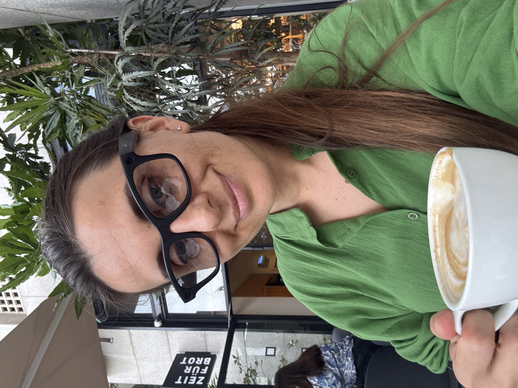 A woman wearing a green shirt and black glasses is holding a white mug with a foamy drink. She is outdoors, possibly at a cafe. In the background, there is greenery, a building with glass doors, and a sign in German that reads "Zeit für Brot." The sign indicates the location is likely a branch of the German bakery chain "Zeit für Brot," which has a location in Tel Aviv, Israel.
