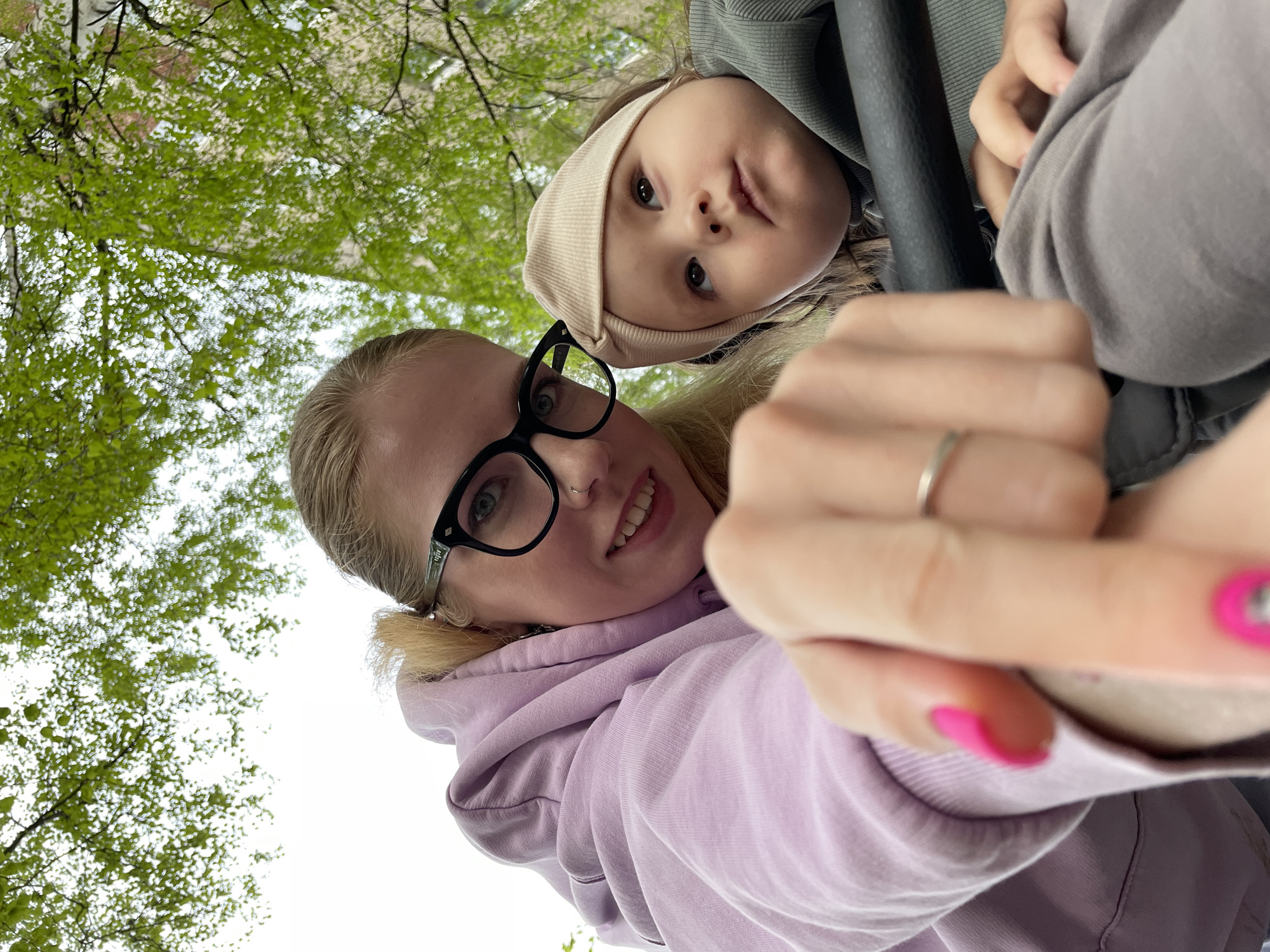 A selfie taken from a low angle features two individuals and a background of trees. The primary subject is a fair-skinned woman with blond hair pulled back into a low ponytail, wearing black-rimmed glasses and a lilac-colored hoodie. She is smiling, and her right hand is raised towards the camera, displaying a silver ring and pink nail polish. Above and slightly behind her is a child with fair skin, wearing a tan-colored head covering and a greyish-green jacket. The child is looking down towards the camera. A dark grey or black vertical bar is present near the child. The background consists of trees with green leaves, suggesting a park or outdoor setting.
The image's location is not discernible as Izhevsk, Russia, based solely on the visual content provided.