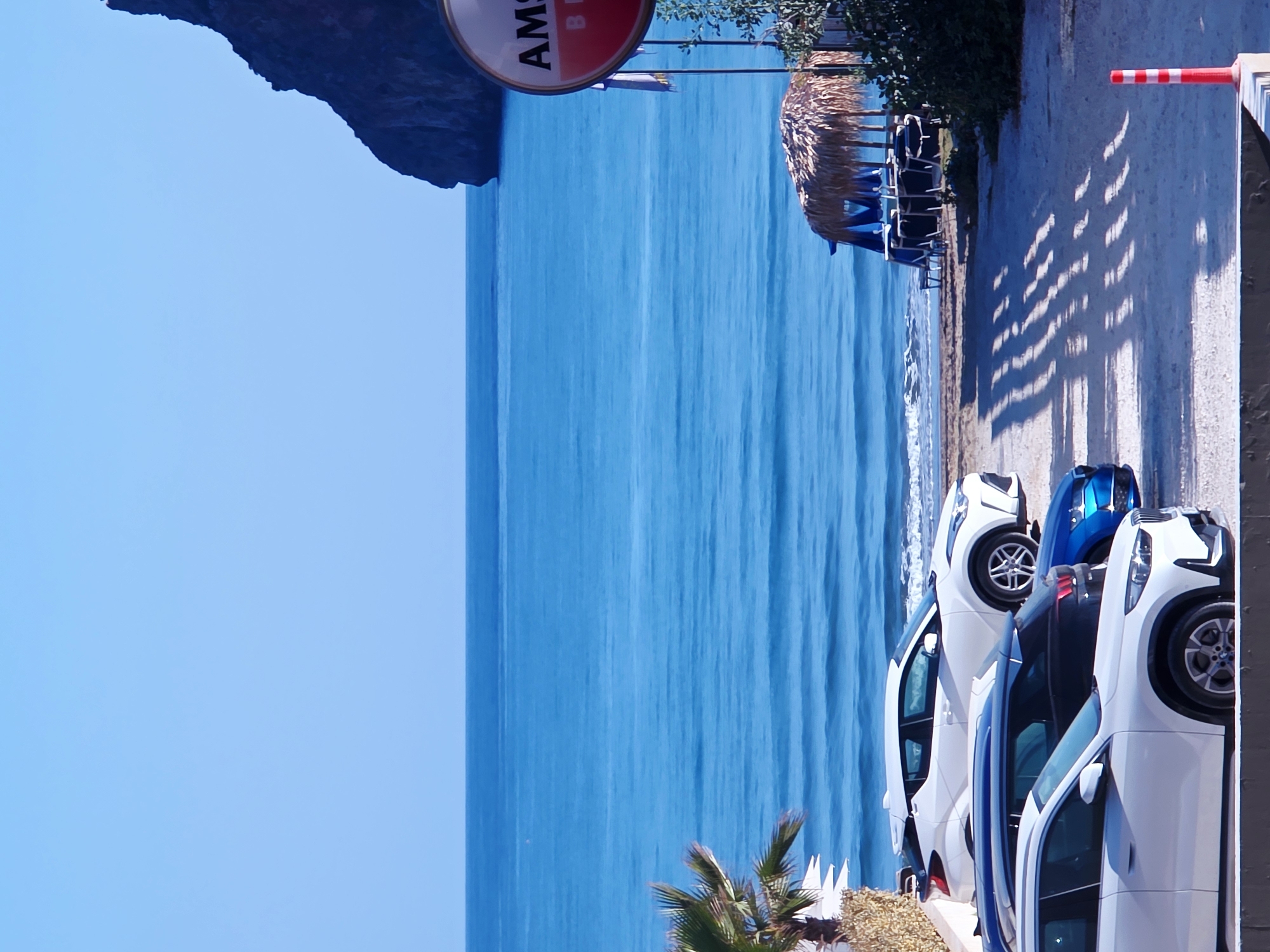 The image depicts a coastal scene in Agía Marína, Greece, with a clear blue sky and the deep blue sea dominating the background. The sea exhibits gentle waves and extends to the horizon. A dark, rocky cliff face is visible on the right side of the frame, partially obscuring the sky. In the foreground, a line of parked cars is visible on a sandy or gravel surface. These include a white SUV, a dark-colored hatchback, a blue sedan, and another white SUV closer to the viewer. Beach chairs and umbrellas, some covered with thatched roofs, are set up on the sandy beach, suggesting a resort or a public beach area. A palm tree with green fronds is present on the left side of the image. A circular sign with red and white lettering, possibly advertising "AMSTEL BEER," is partially visible on a pole on the right. A striped bollard stands in the lower right corner. The overall impression is a sunny day at a Mediterranean beach.