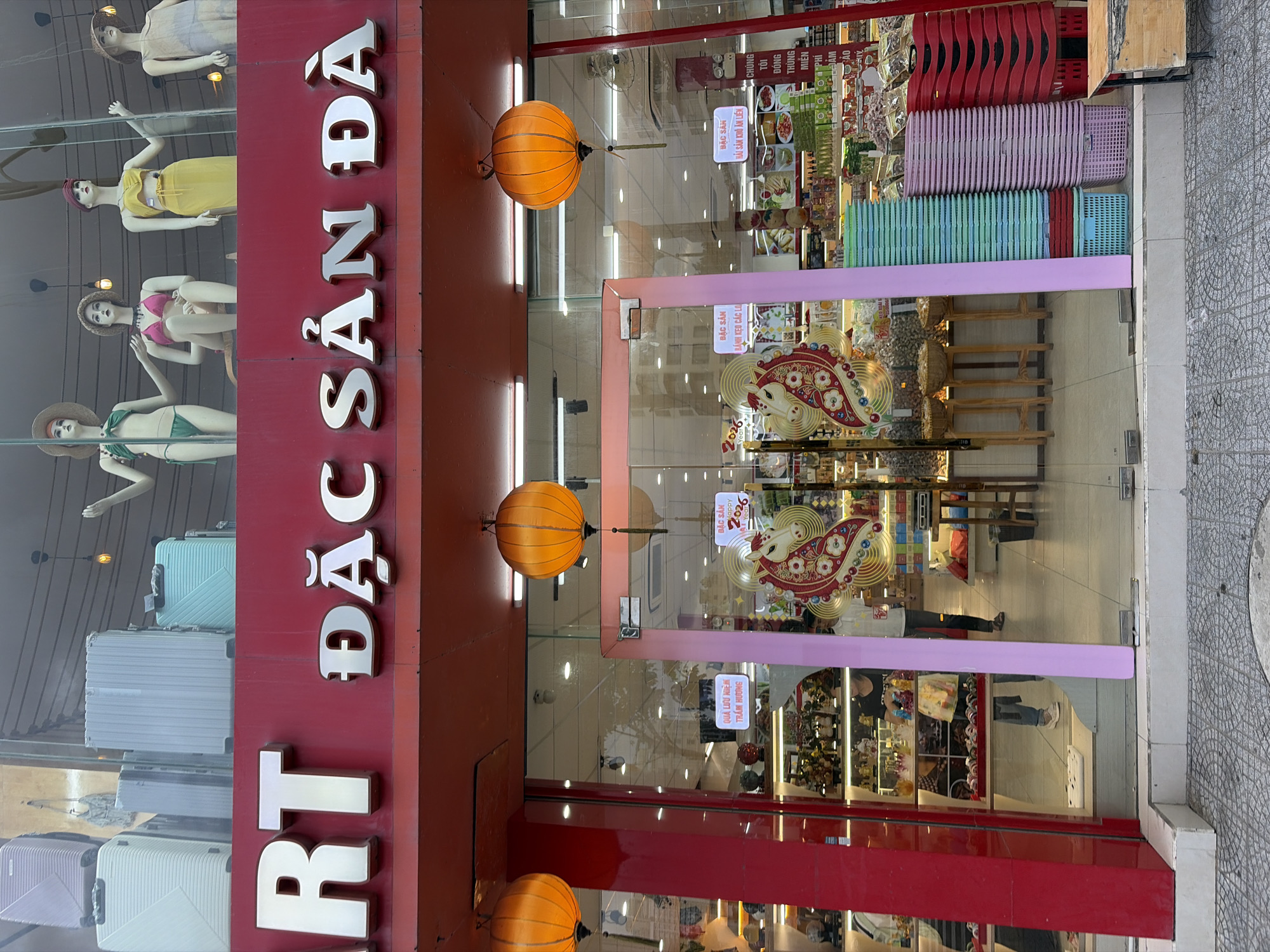 The image shows the storefront of "MART ĐẶC SẢN ĐÀ NẴNG", a specialty store in Da Nang, Vietnam. The store features a bright red facade with large, silver, 3D letters spelling out the store's name. The upper portion of the storefront displays mannequins dressed in swimwear and hats, suggesting the sale of beachwear or resort clothing. Below this, the main display area is visible through large glass windows, showcasing a variety of products arranged on shelves. Ornate orange lanterns hang from the ceiling above the entrance, adding a festive atmosphere. In the foreground, a doorway with pink trim leads into the store. To the right of the entrance, stacks of colorful plastic baskets, primarily green, pink, and red, are neatly arranged, indicating their availability for purchase or use within the store. A small wooden table with a metal frame is positioned outside the entrance on the right. The street in front of the store is paved with patterned tiles.