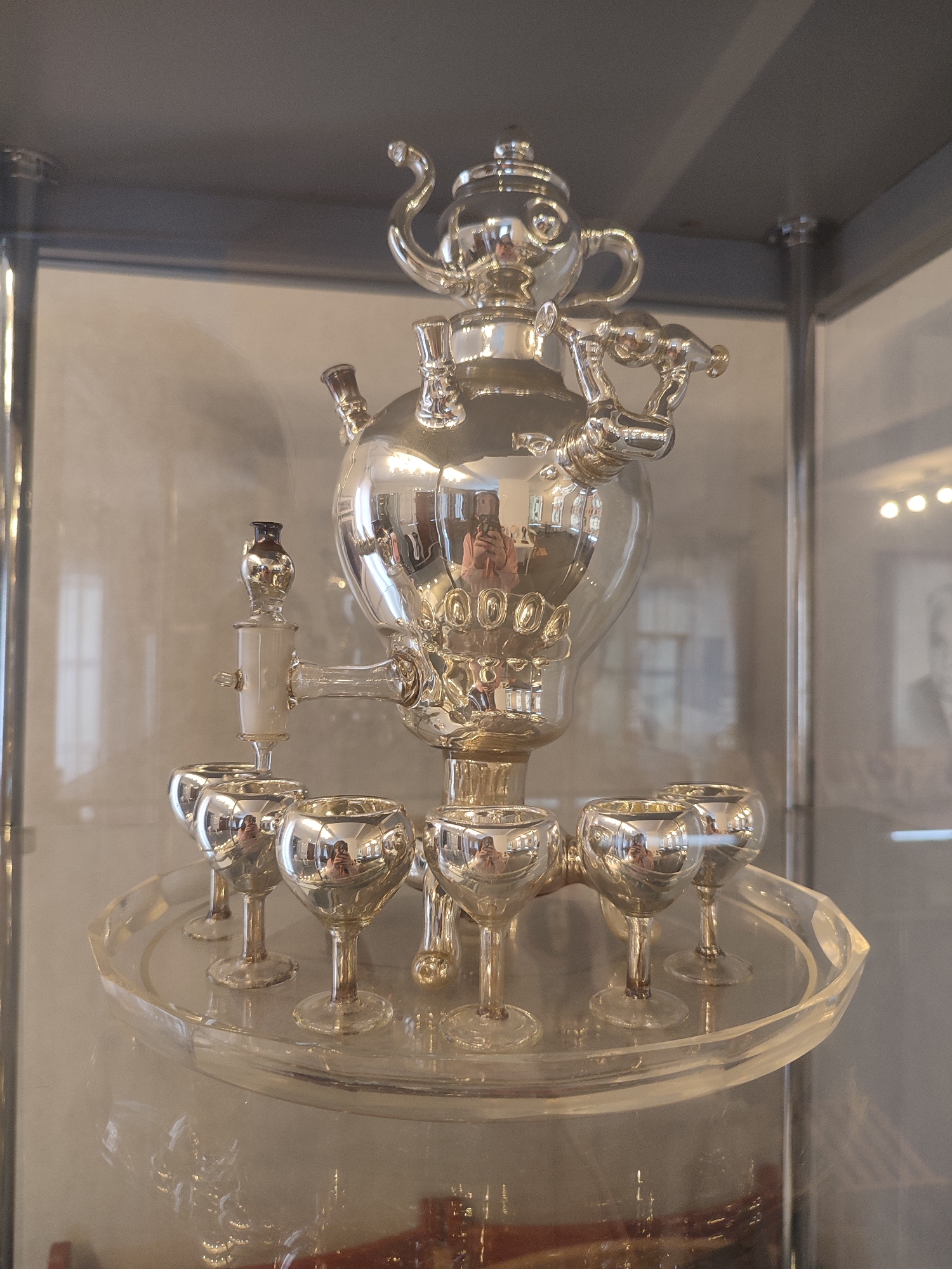 A silver samovar and cordial glasses are displayed on a glass stand inside a glass display case. The location is the Sarapul History and Culture Museum in Sarapul, Russia. The reflections in the metal surfaces reveal interior lighting and a person holding a camera.
