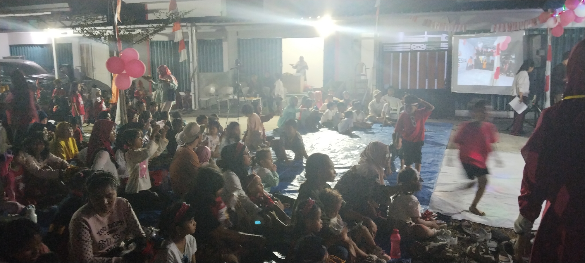 The image depicts an outdoor gathering in what appears to be a residential or community area in Taban, Indonesia. A large group of people, predominantly children, are seated on the ground covered by a blue tarp, facing a projection screen. The screen displays an image of what seems to be a group of people in formal attire, possibly at an event. Balloons, some pink, are visible around the area, along with Indonesian flags, suggesting a celebratory occasion. A tripod with a camera is set up near the screen, indicating that the event is being filmed. A person in a red shirt and dark shorts is walking on a white sheet in the foreground, while another person in a dark red robe stands to the right, partially obscuring the view. The overall atmosphere suggests a community event or celebration, possibly a children's gathering or a public viewing of a recorded event.
