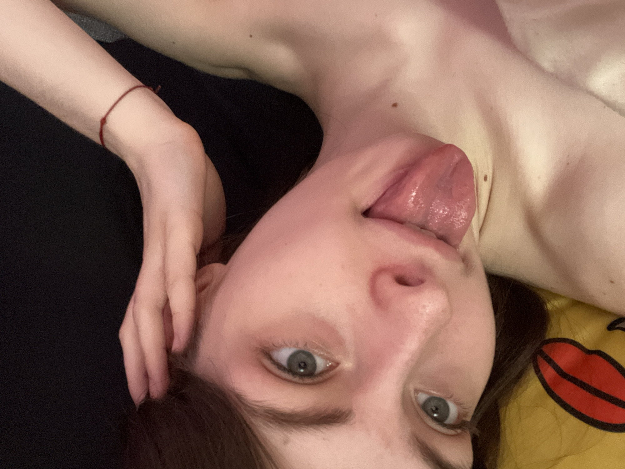 A close-up, upside-down view features a light-skinned person with brown hair and grey eyes sticking out their tongue. A thin red bracelet is visible on their left wrist. The person is lying on what appears to be a yellow pillow with a red lip design. The background includes dark fabric. The location is unknown, Russia.