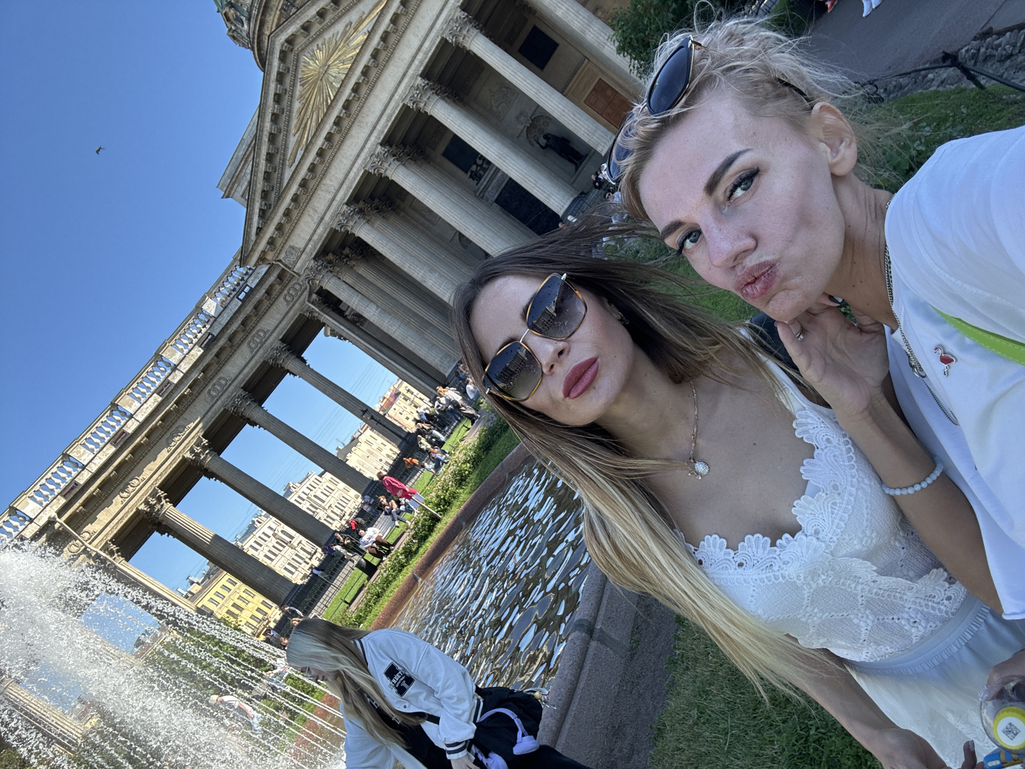 This is a selfie taken outdoors with a focus on two women in the foreground. The woman on the right, with blonde hair pulled back and sunglasses perched on her head, is puckering her lips as if for a kiss. She wears a white top adorned with a small flamingo pin. Next to her, a woman with long blonde hair and stylish sunglasses gazes at the camera, her reflection visible in her sunglasses. She is wearing a white lace top. In the background, a grand building with towering columns and an ornate facade is visible. A fountain sprays water in the lower left corner of the image, adding a refreshing element to the scene. The sky is a clear, bright blue, suggesting a sunny day. The presence of other people in the background, some sitting and others standing, indicates a public space, possibly a park or a plaza. The architecture suggests the location is Saint Petersburg, Russia, perhaps Kazan Cathedral given the architectural style. The overall mood appears cheerful and relaxed, characteristic of a tourist outing or a casual meet-up. No visible text is discernible in the image.