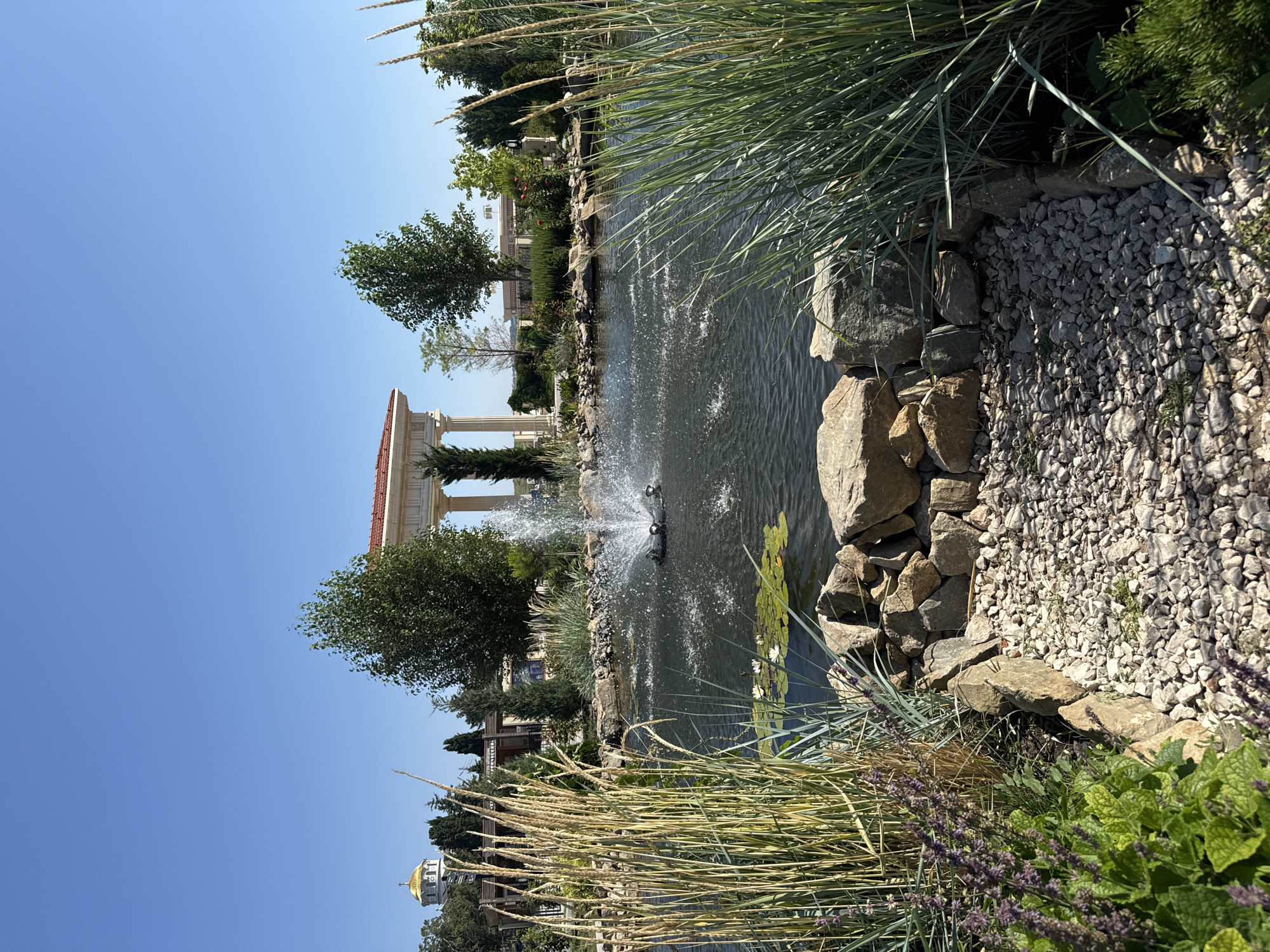 A photograph captures a serene outdoor scene with a pond and architectural elements. The pond is the central focus, featuring a black fountain device that sprays water upwards, creating a dynamic splash. Lily pads, some with white flowers, float on the surface of the dark water. The pond's edge is adorned with a rock border, filled with light-colored gravel on the right side of the image. Tall, dry grasses and vibrant green plants frame the foreground, with purple flowers blooming in the lower left.
In the background, behind the pond, a classical-style colonnade with a red-tiled roof stands prominently. To the left of the colonnade, a white building with a golden dome, resembling a church or a similar religious structure, is visible. Several trees with green foliage are scattered throughout the background, adding to the natural beauty of the setting. The sky is a clear, bright blue, indicating a sunny day. The overall impression is one of tranquility and picturesque landscaping. The location appears to be the Chersonesus National Historic and Archaeological Museum-Reserve in Sevastopol, Ukraine, a UNESCO World Heritage site known for its ancient Greek ruins and beautiful grounds.