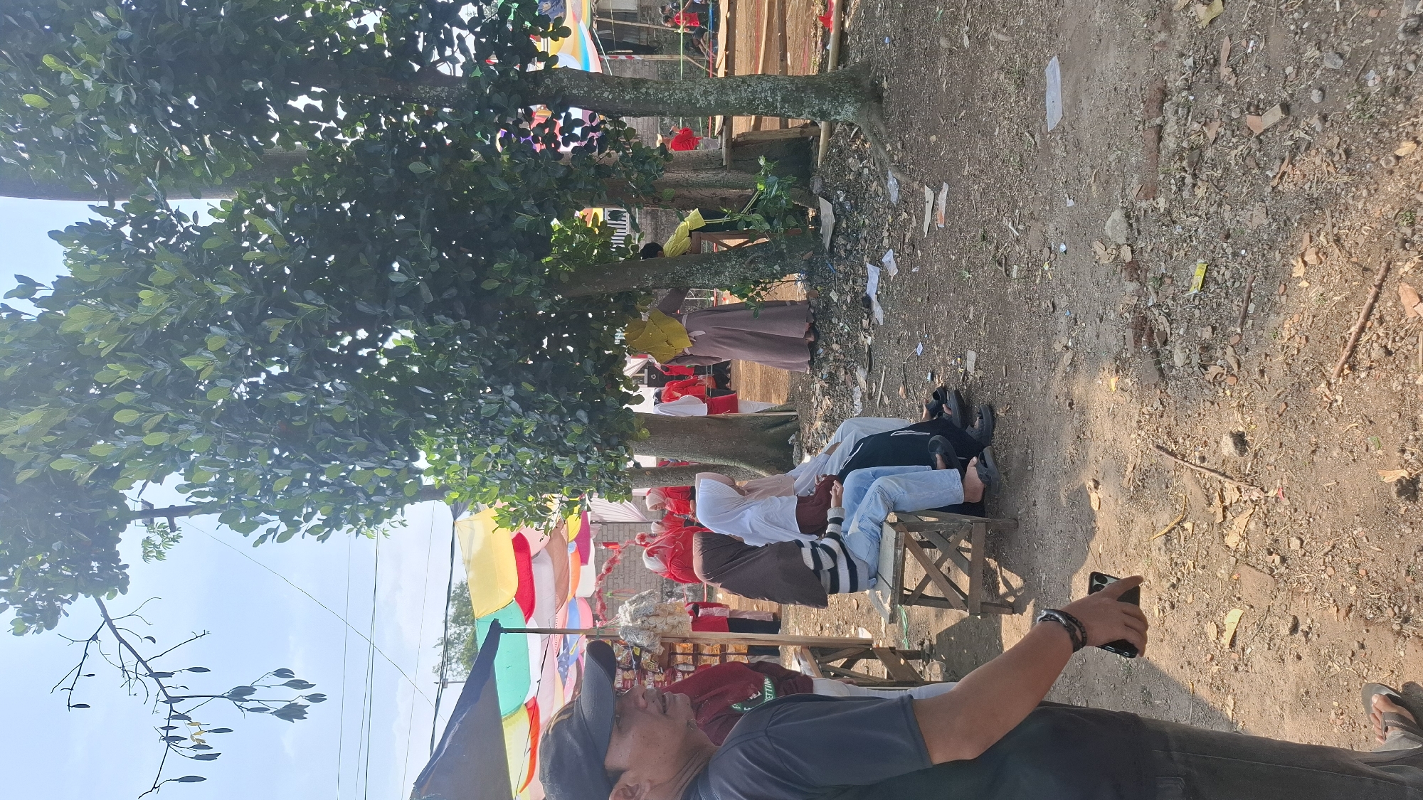 The image shows an outdoor gathering, possibly a community event, in Cilaku, Indonesia. In the foreground, a man wearing a black t-shirt and a black baseball cap is looking to the left, holding a dark-colored smartphone in his right hand. Behind him, two women are seated on a wooden bench. The woman on the left wears a brown headscarf and a striped shirt with blue jeans, while the woman on the right wears a white headscarf and a white top with jeans.
In the middle ground, several individuals are visible in the background, some wearing red attire and headscarves, suggesting a cultural or religious event. Colorful canopies in red, yellow, and white are stretched overhead, providing shade. The environment appears to be a dirt-covered area with scattered debris, including leaves and small pieces of wood. Trees with dense green foliage are prominent throughout the scene, casting shadows on the ground. The sky is bright blue with a few scattered clouds. Further in the background, there are hints of structures and possibly more people gathered. The overall impression is of a lively, informal event taking place during daylight.