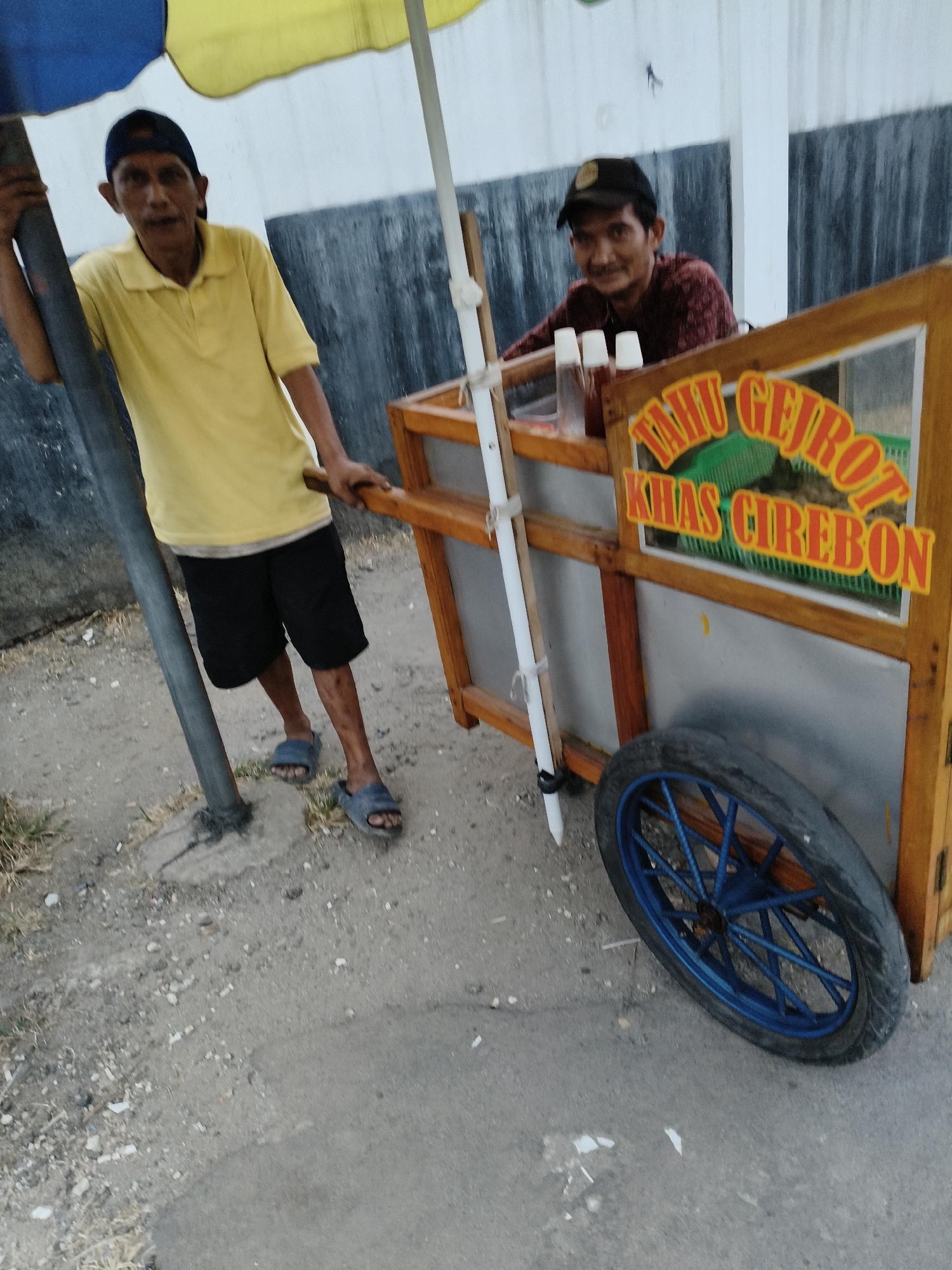 In the city of South Tangerang, Indonesia, on a grey, rough, sandy street, two men are standing beside a food cart. The cart, made of wood and metal, is equipped with a wheel with blue spokes and a sign in Indonesian that reads "TAHU GEJROT KHAS CIREBON." One man, wearing a yellow t-shirt and black shorts, stands to the left of the cart, holding onto a grey pole. He appears to be of elderly age, with weathered skin and a visible mustache. The second man, younger, with a dark cap and a maroon patterned shirt, is positioned behind the cart, facing the camera with a slight smile. He is holding up two white cups. A yellow and blue umbrella is extended above the food cart, providing shade. The background consists of a textured grey wall. The lighting suggests it is either early morning or late afternoon.