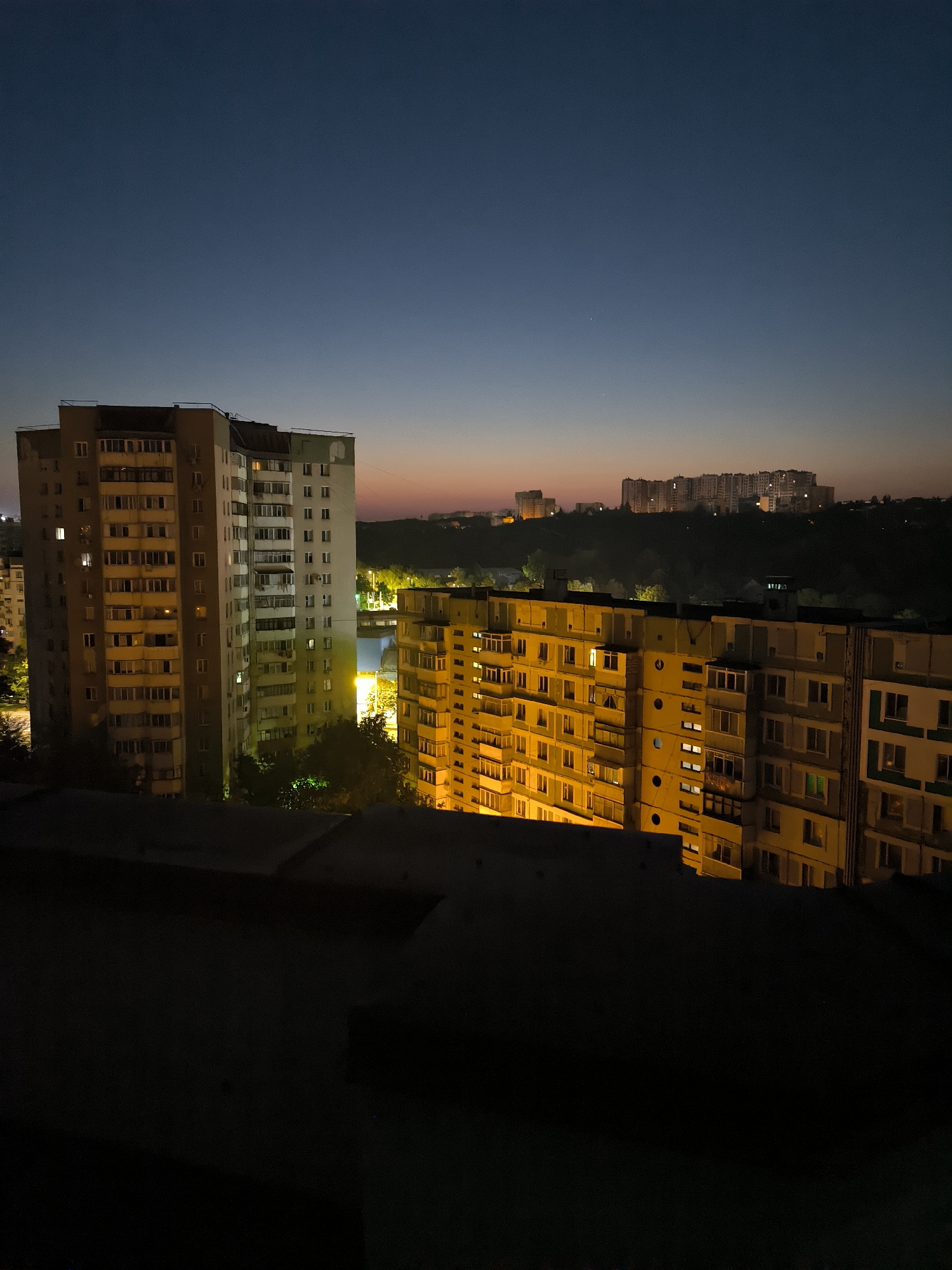 The image depicts an urban skyline at dusk or dawn, with several apartment buildings illuminated by interior lights. The sky is a gradient of deep blue at the top, transitioning to a lighter blue and then a faint orange hue near the horizon. In the foreground, a dark, silhouetted rooftop or balcony railing occupies the bottom portion of the frame.
To the left, a tall, light-colored apartment building with a rounded edge stands out. It has numerous balconies and windows, many of which are lit, suggesting occupancy. To the right, another, shorter apartment building is visible, predominantly lit with a warm, yellow light emanating from its windows. This building also features many balconies and windows.
In the background, a series of larger, more distant apartment buildings are visible, spread across a dark, tree-covered hillside. These buildings are also illuminated, creating a scattered pattern of lights against the darkening landscape. The overall impression is one of a residential area settling into or waking from nighttime.
While the image is a general urban landscape and does not contain specific identifiable landmarks that would pinpoint its exact location within Chișinău, Moldova, the architectural style of the apartment blocks, particularly the older, pre-fabricated panel buildings with their characteristic balconies, is consistent with residential areas found in Chișinău and other cities in Eastern Europe. The presence of dense, high-rise apartment buildings forming the skyline is also characteristic of many Soviet-era urban developments.