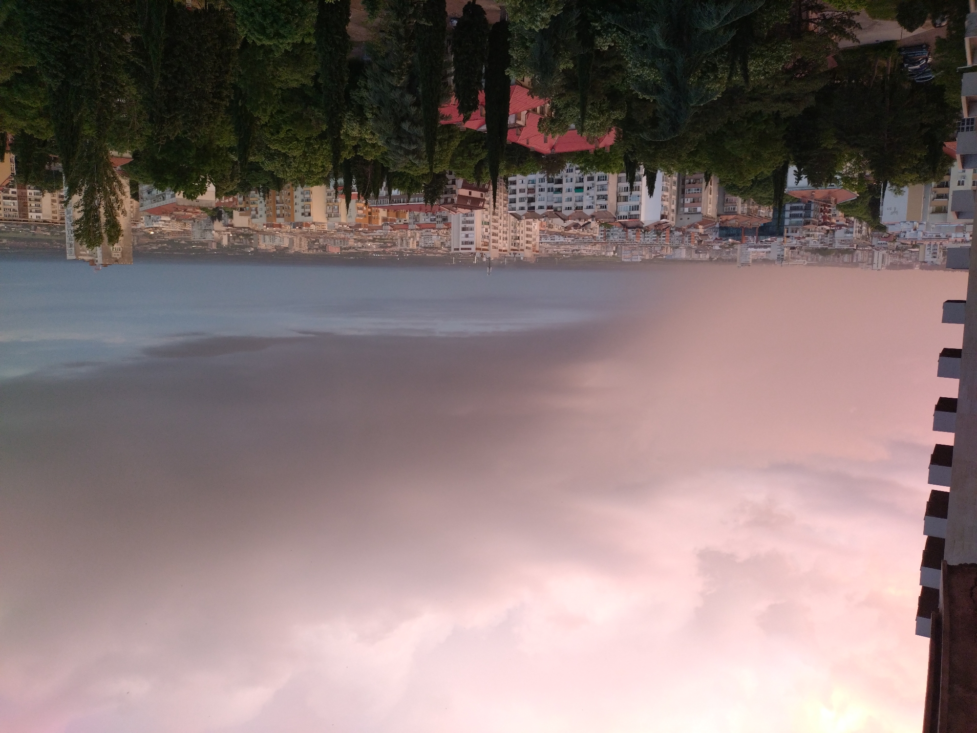 The image depicts a cityscape of Stara Zagora, Bulgaria, viewed upside down. The upper portion features the urban landscape, comprising residential and commercial buildings, dense trees, and a body of water that appears calm. Below, the sky is filled with pink and gray clouds, giving the impression of either sunrise or sunset. On the right side of the frame, a building facade is visible, hinting at the perspective of an elevated location. The photograph is oriented in such a way that the sky dominates the foreground while the city becomes part of the upper background.