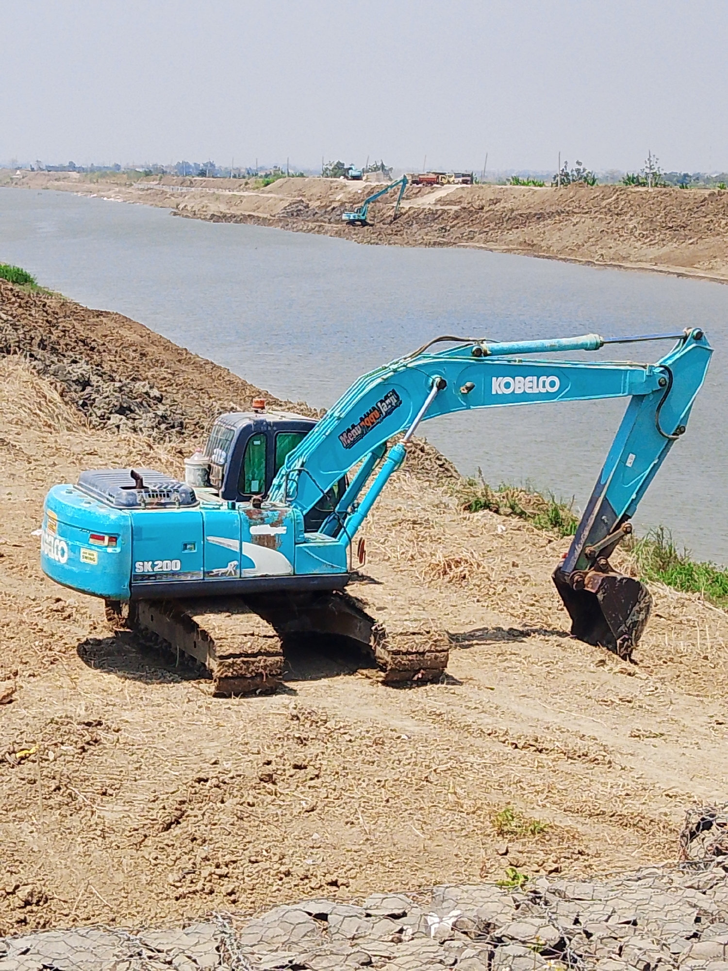 The image depicts a construction site near a body of water, likely a canal or river, in Bengkalis, Indonesia. A large blue Kobelco SK200 excavator is positioned on a dirt embankment, with its tracks digging into the dry earth. The excavator is a prominent subject, showing signs of use with mud and wear on its body and bucket. In the background, the hazy sky suggests a warm, possibly humid climate typical of the region. The landscape is characterized by earthen mounds and a wide expanse of water. Several other construction vehicles, including trucks and what appear to be smaller excavators, are visible in the distance on the opposite bank, indicating ongoing large-scale earthmoving operations. The overall scene suggests a significant infrastructure or development project underway in a rural or semi-rural setting within Bengkalis.