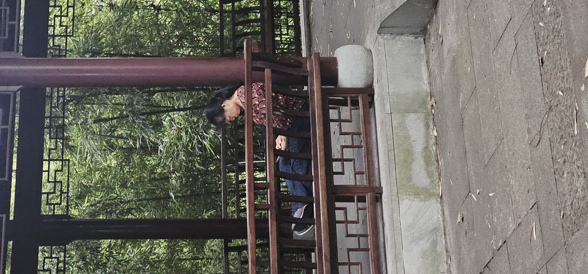 Captured in Yuzhong District, Chongqing, China, the image shows a woman within the confines of a traditional Chinese structure. She is wearing a patterned red and black long-sleeved top paired with denim jeans and athletic shoes. Her head is bowed, and her hands are clasped in front of her. The structure is made of dark brown wood with a complex geometric pattern, possibly part of a pavilion or gazebo. Lush green bamboo foliage is visible in the background, creating a serene backdrop. The immediate foreground features a concrete curb adjacent to a stone-paved pathway.