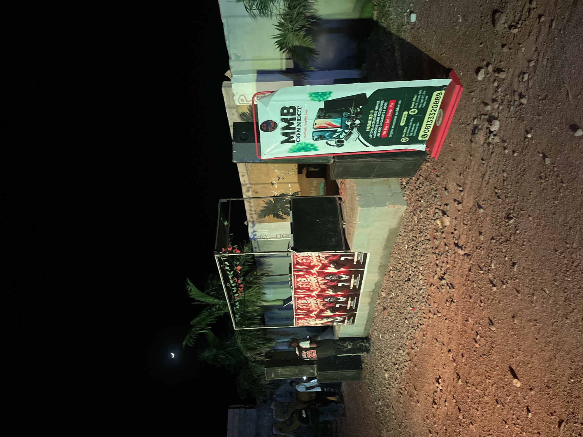 The image depicts an outdoor event scene at night in Durumi, Nigeria. A DJ booth is set up with speakers and a laptop on a raised platform. Several posters are displayed on the front of the DJ booth. A banner for "MMB CONNECT" stands to the right of the DJ booth, advertising services related to electronic devices. The ground is covered with dirt and gravel, and a wall runs along the background. A crescent moon is visible in the dark sky, along with some palm trees. Several individuals are present, some appearing to be staff or attendees. One person in the foreground is wearing a black vest with the number "23" on the back and appears to be looking at their phone. The overall atmosphere suggests a local gathering or party.