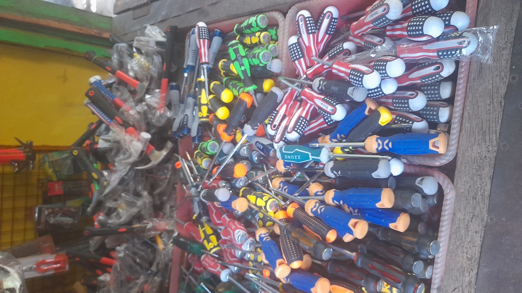 The image shows a close-up, overhead view of a collection of screwdrivers and other tools displayed in a wooden bin. The tools are a mix of various sizes and colors, with some featuring red, blue, yellow, and green handles. Many of the screwdrivers have plastic packaging. In the background, there are more tools visible, including hammers and toolkits, some of which are also in plastic packaging. The overall setting appears to be an outdoor market stall. The yellow wall and wooden structures suggest a location in Pekalongan, Indonesia, likely a hardware or tool vendor. No people are visible in the frame.