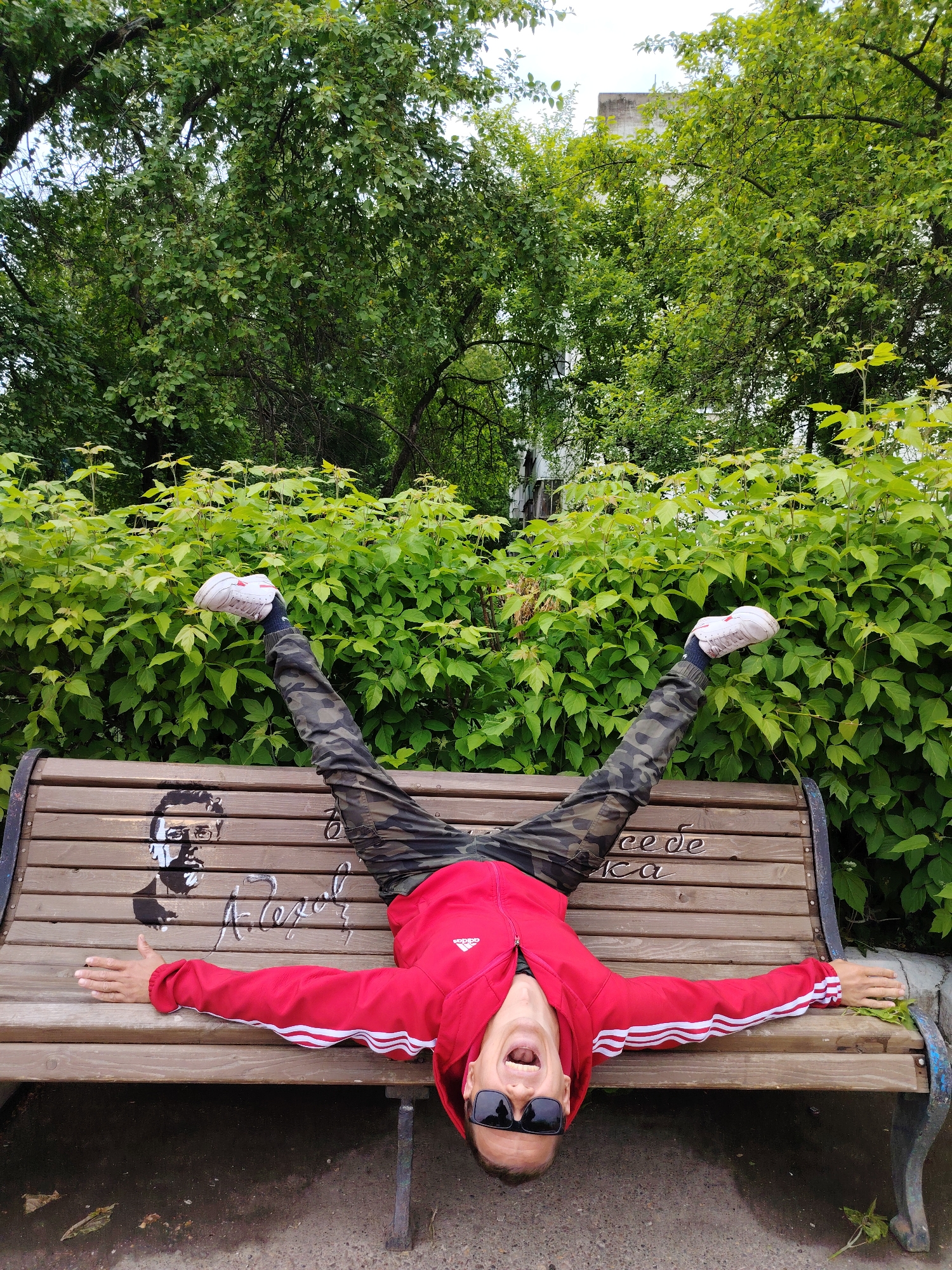 A person lies upside down on a park bench in Chaikovsky, Russia. The individual, wearing sunglasses, a red Adidas track jacket, and camouflage pants, has their arms outstretched along the bench and legs raised upwards, spread in a V-shape. They are wearing white sneakers. The person's mouth is open, appearing to shout. The bench is wooden with writing and a drawing on it, and the surrounding environment consists of trees, bushes, and foliage typical of a park setting. The ground beneath the bench is paved.