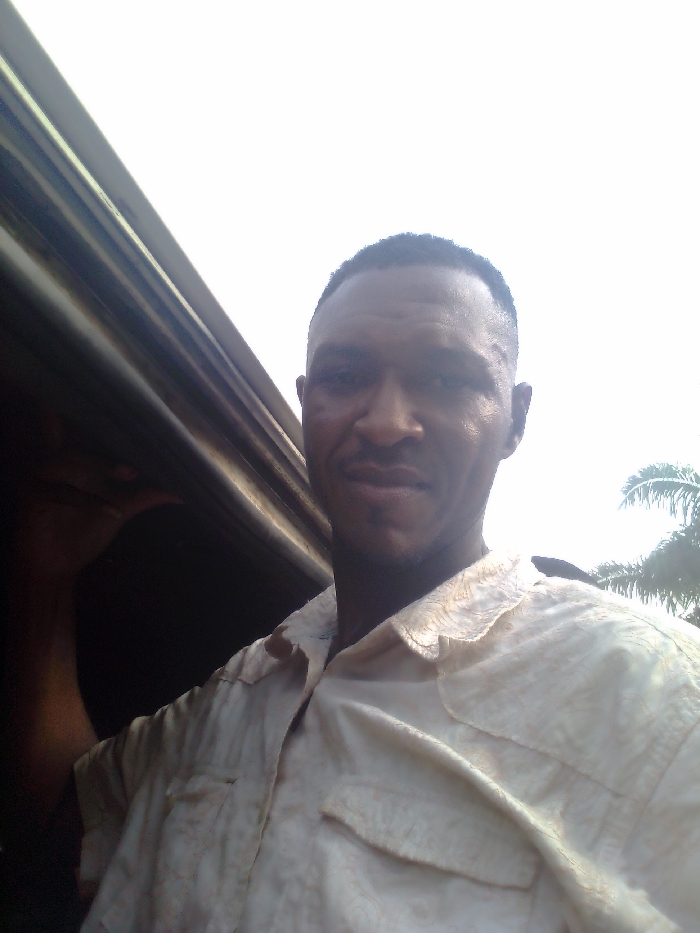 A man in a pale, button-down shirt stands beside a dark, metallic surface, possibly a car or vehicle door. His expression is neutral. Palm trees are visible in the background, hinting at a tropical or subtropical location. The lighting is bright and somewhat diffuse. He has short hair and appears to be of African descent. The overall composition is informal and snapshot-like.