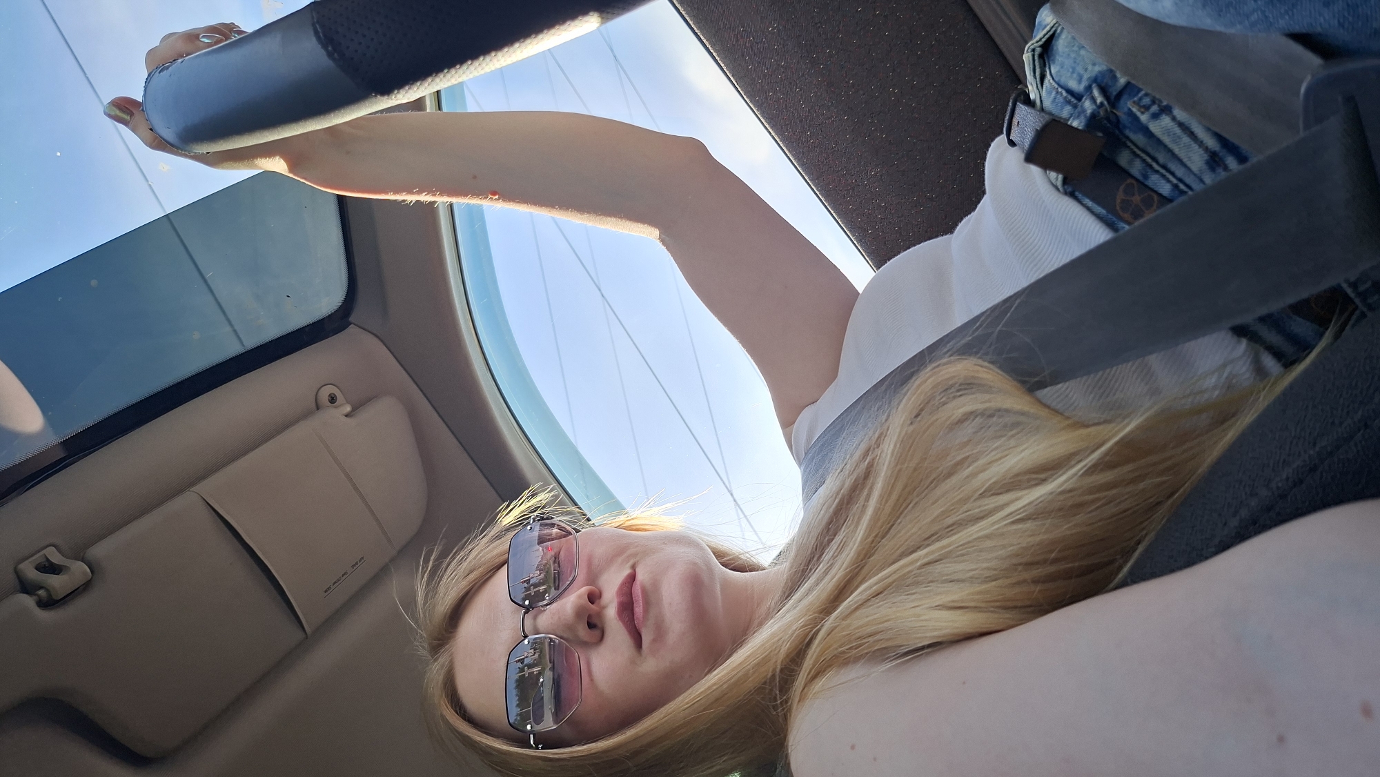 A woman with long blonde hair and sunglasses is seated in a car, secured by a seatbelt. The car has a sunroof, through which the sky and power lines are visible. The car's interior includes a sun visor. The reflection in the sunglasses reveals urban surroundings, including a building and other vehicles. Based on the prompt, the location is Chaikovsky, Russia.