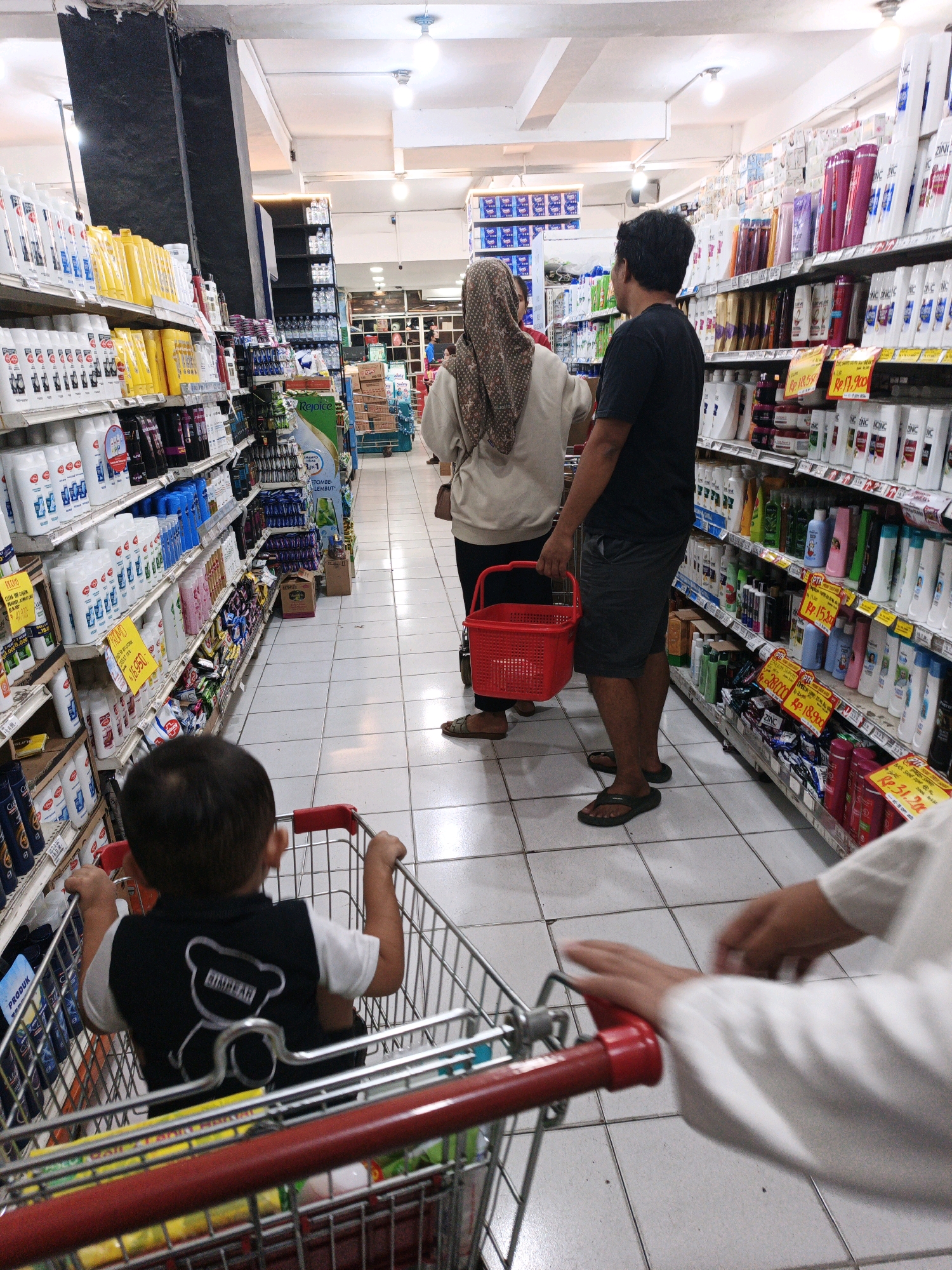 In the city of Jagalan, Indonesia, a family navigates a brightly lit supermarket aisle stocked with hygiene products. A young boy, seated in a shopping cart, faces away from the camera, his hands gripping the cart's handles. His black vest features a white teddy bear graphic with the word "BUMBLE" underneath. To his right, a woman wearing a patterned hijab and a light-colored shirt stands beside a man in a black t-shirt and shorts. The man holds a red shopping basket. The tiled floor is clean, and shelves laden with various shampoos, conditioners, and lotions line both sides of the aisle. The scene is captured from a low angle, emphasizing the child's perspective within the shopping cart.