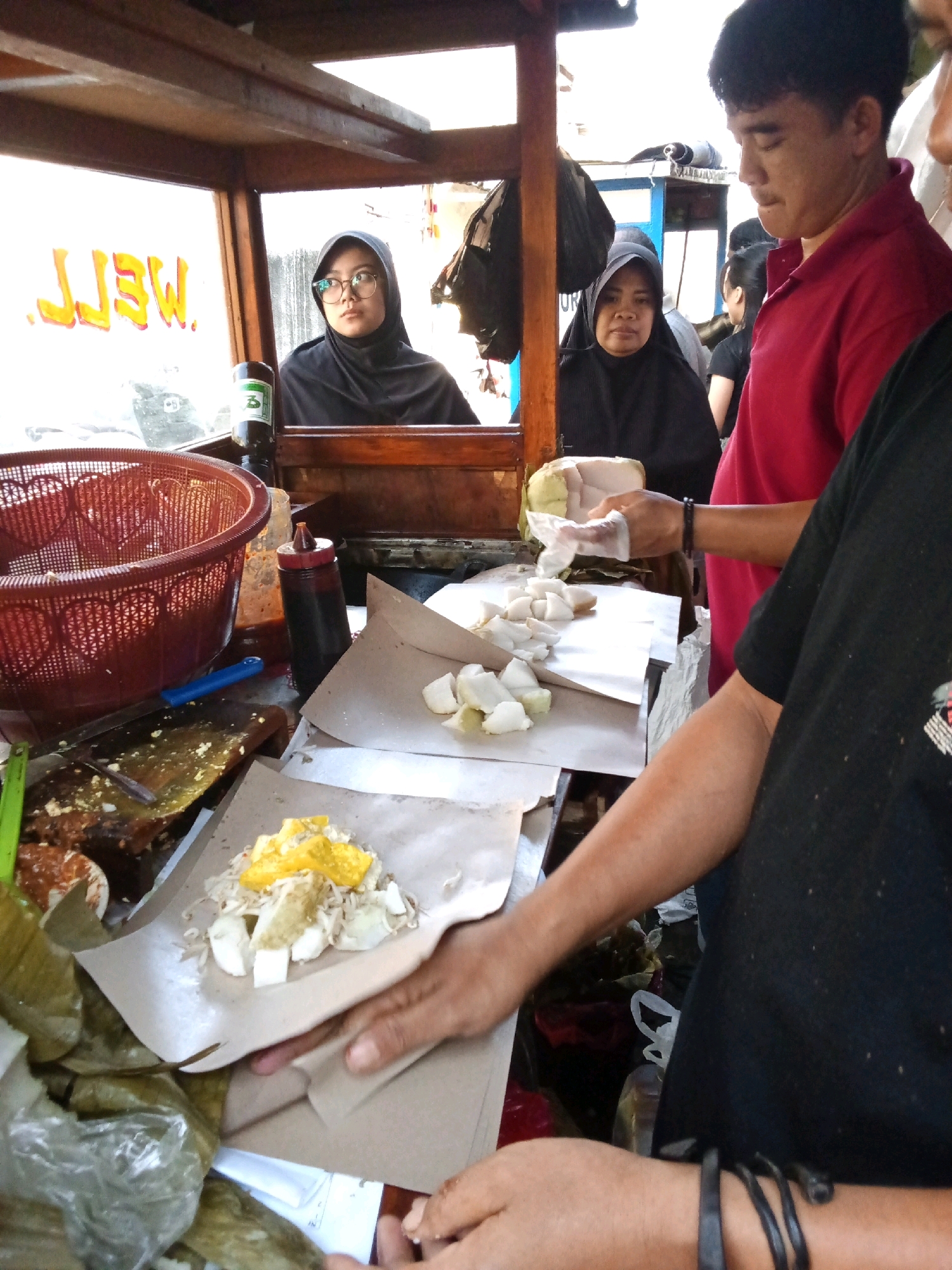 In the city of Bandung, Indonesia, this photograph captures a street food vendor preparing and serving food. The scene is set at a food stall, likely a mobile cart, with a wooden frame and a clear plastic or glass front. Two women, both wearing hijabs, are visible behind the counter. One woman on the left, wearing glasses, appears to be looking to the side. The other woman, in the center, is looking forward. A man in a red shirt stands to the right, looking down and to the left, engaged in the food preparation. Another person, wearing a black shirt, is partially visible on the far right, with their arm extended towards the counter.
On the counter, various food items are laid out on paper or banana leaves. There are chopped white ingredients that resemble tofu or rice cakes, and some sprouts. One portion of food is topped with yellow pieces, possibly egg or cheese. A red basket, a blue utensil, and bottles of sauce are also visible. The background shows more food items and the interior of the stall. The lighting suggests it is daytime, with natural light illuminating the scene. The overall impression is a busy street food vendor serving customers.