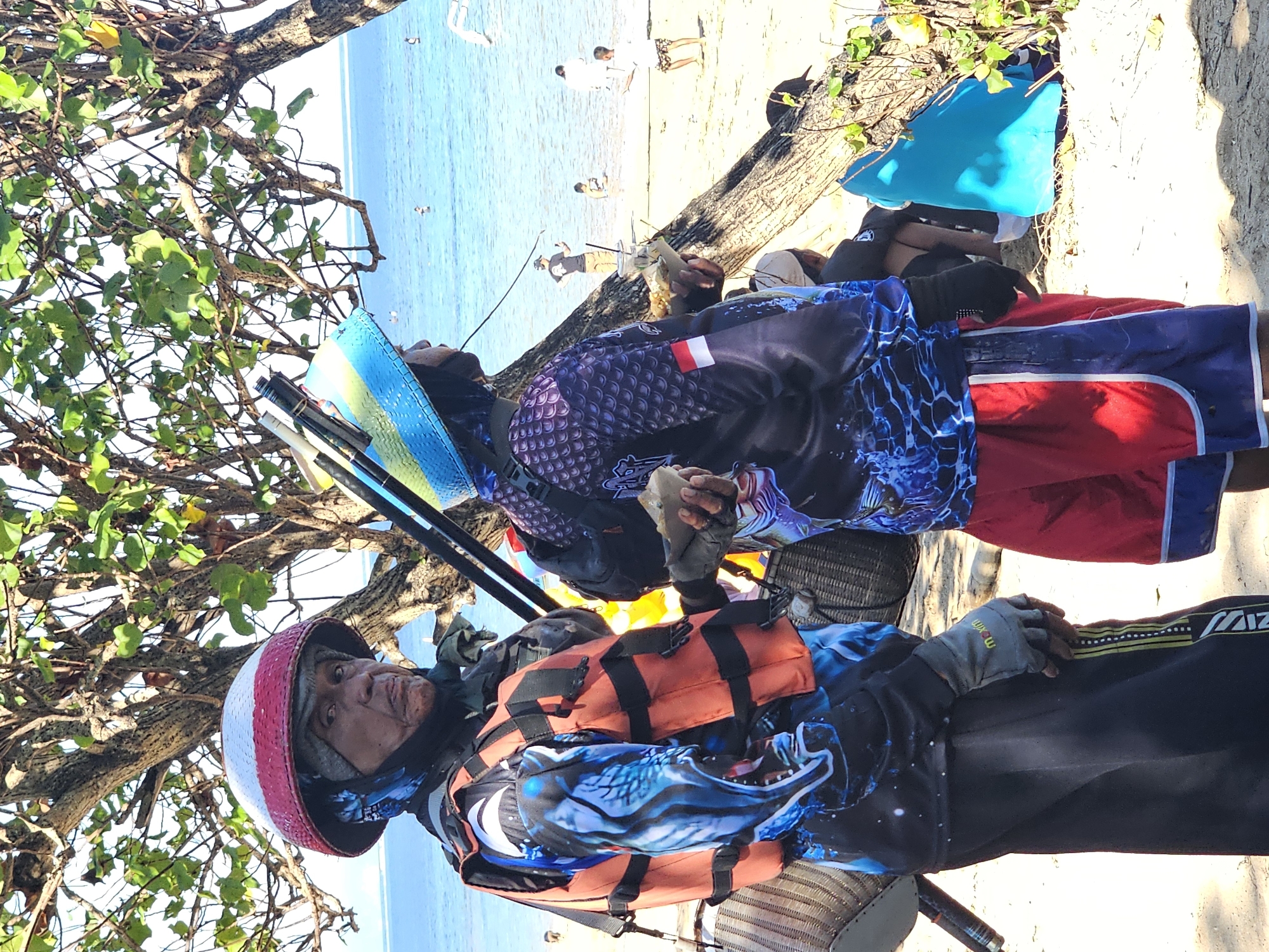 Two individuals, likely fishermen, stand on a sandy beach in Denpasar, Indonesia, near the ocean. The person on the left, an older male wearing a red and white conical helmet and an orange life vest over a blue and black long-sleeved shirt with a graphic print, faces the camera. He wears black pants with a striped side detail and gray gloves. Behind him, leaning against a tree, is a woven basket and fishing gear. To the right, a second individual, wearing a blue and green conical helmet, is seen from the side, also in a long-sleeved shirt with a graphic print and red shorts. Both individuals appear to be holding food items wrapped in paper. The background shows the turquoise ocean with a few people wading in the water and distant landmass under a bright, sunny sky. The scene suggests a casual, possibly recreational, fishing activity on a tropical coastline.