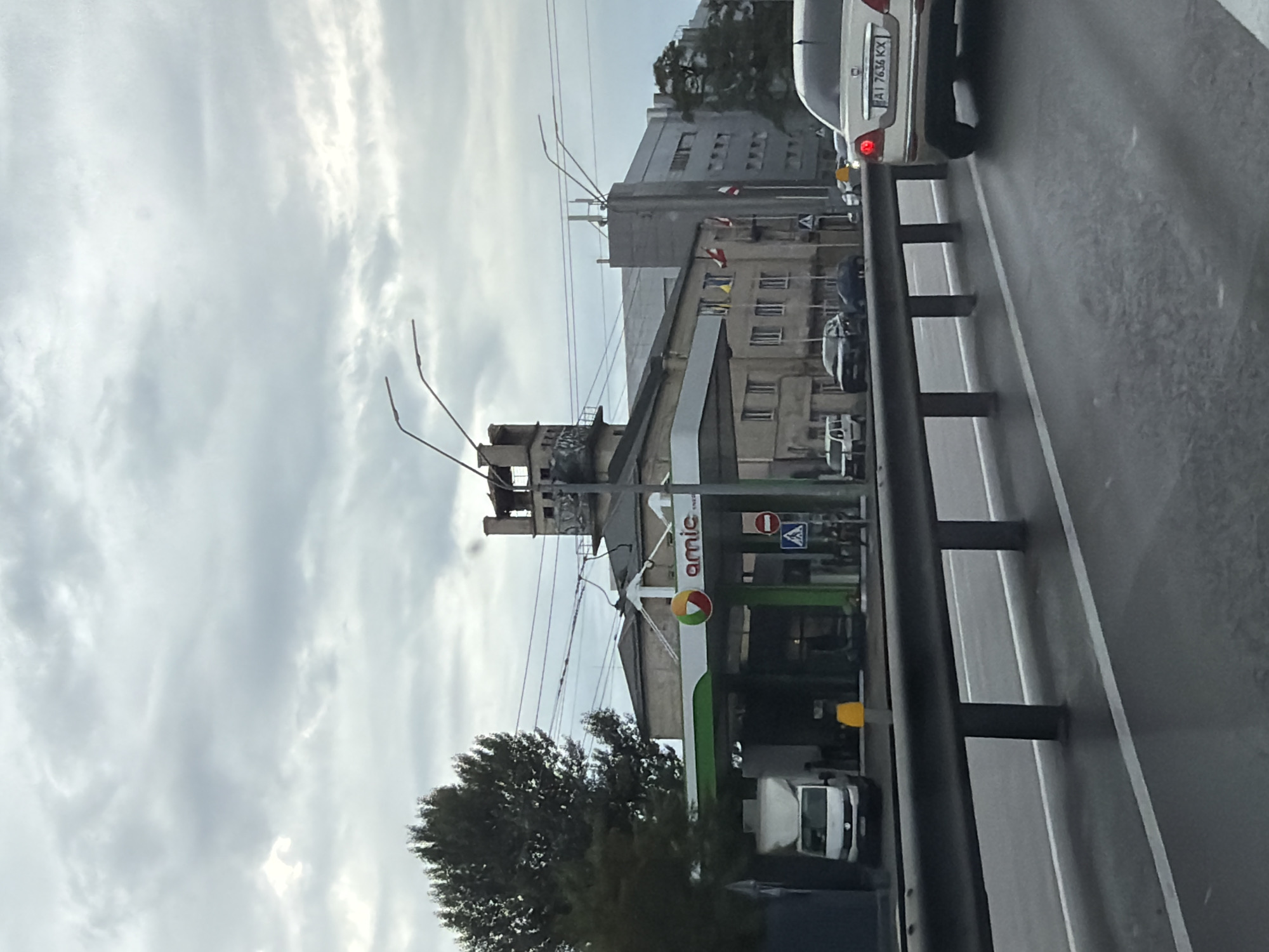 The image shows a view from a moving vehicle on a cloudy day in Kyiv, Ukraine. In the foreground, a dark metal guardrail separates the road from the shoulder. Beyond the guardrail, a street with wet asphalt and white lane markings is visible. Several cars are on the road, including a light-colored sedan with license plate AI 7636 KX in the right lane.
In the middle ground, a gas station with the "amic" logo is prominent. There are pumps and a white van at the station. Behind the gas station, a multi-story building with windows and flags hanging from poles is visible. To the right of this building is a larger, modern grey building. In the background, trees and power lines can be seen. The sky is overcast with grey clouds.