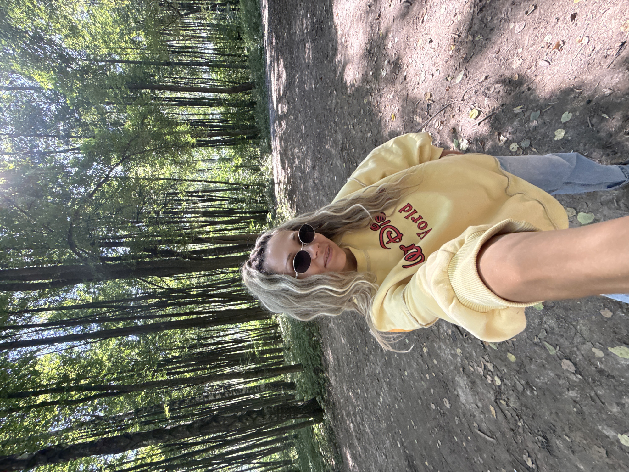 In the city of Rusanovo, Russia, a woman with blonde, curly hair pulled back in a ponytail is taking a selfie in a forest. She is wearing sunglasses and a yellow sweatshirt with red lettering that reads "Disney World." The sweatshirt is adorned with a graphic of Mickey Mouse. She is also wearing blue jeans. The woman is standing on a dirt path, with a dense forest of tall trees surrounding her. Sunlight filters through the leaves, creating dappled shadows on the ground. The path is covered with fallen leaves and small branches.