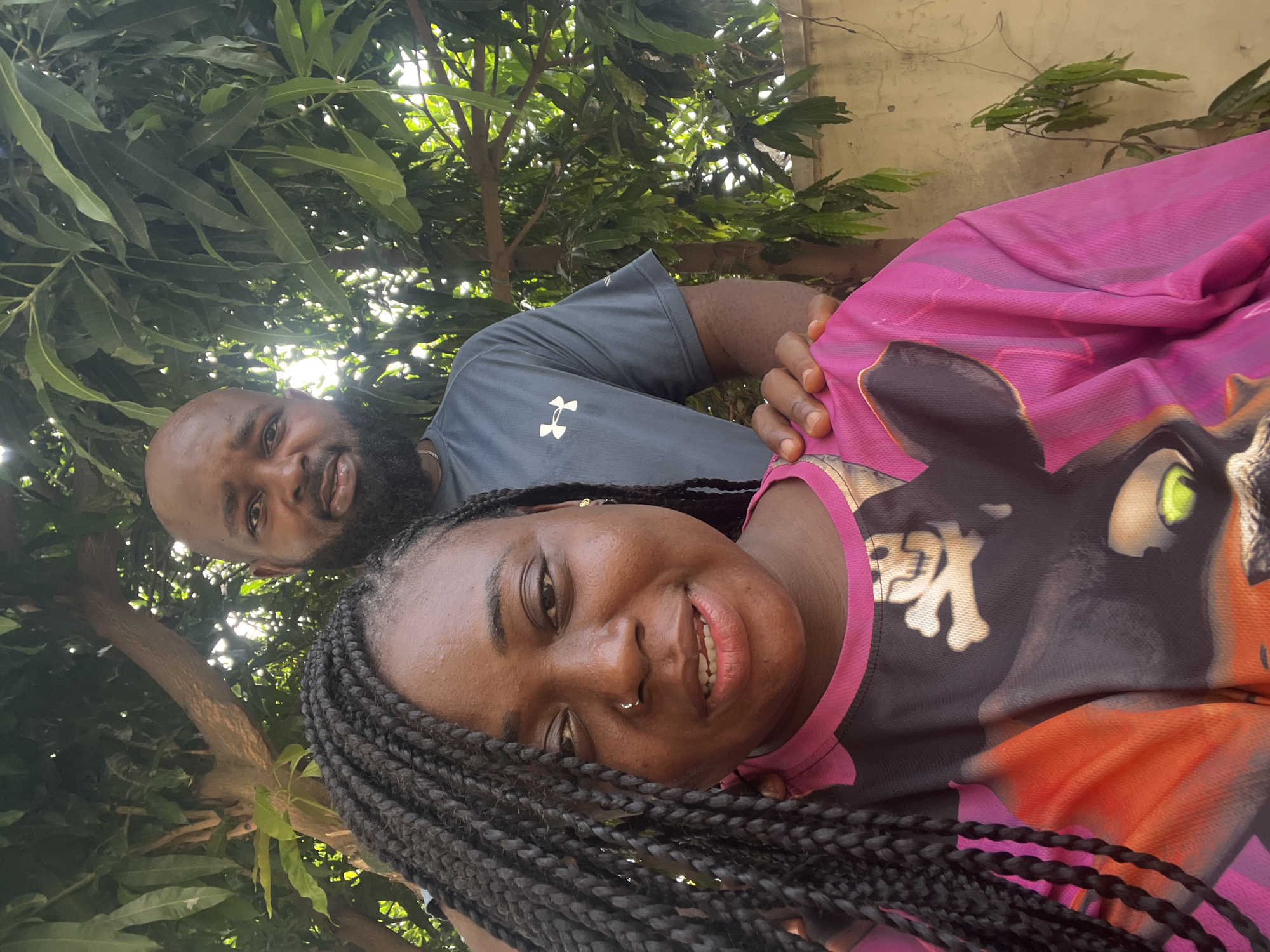 A brightly lit outdoor portrait, likely taken in Kubwa, Nigeria, captures two smiling individuals posing closely together. In the foreground, a woman with dark braided hair and a nose piercing smiles warmly towards the viewer, her teeth slightly visible. She wears a vibrant pink and black top adorned with a cartoonish cat-like creature featuring bright green eyes, alongside a skull and crossbones emblem. A small gold earring is visible on her right ear.
Behind and slightly to her right, a man with a bald head and a full dark beard is partially visible, also smiling. He wears a grey t-shirt that displays the "Under Armour" logo prominently. His left arm is seen resting around the woman's shoulder, suggesting a close relationship.
The background is filled with abundant lush green foliage from trees, indicating a natural outdoor setting, possibly a garden or yard. Bright highlights on the leaves suggest clear daylight. A light-colored wall is partially visible in the upper right background. The overall impression is one of happiness and warmth shared between the two subjects.