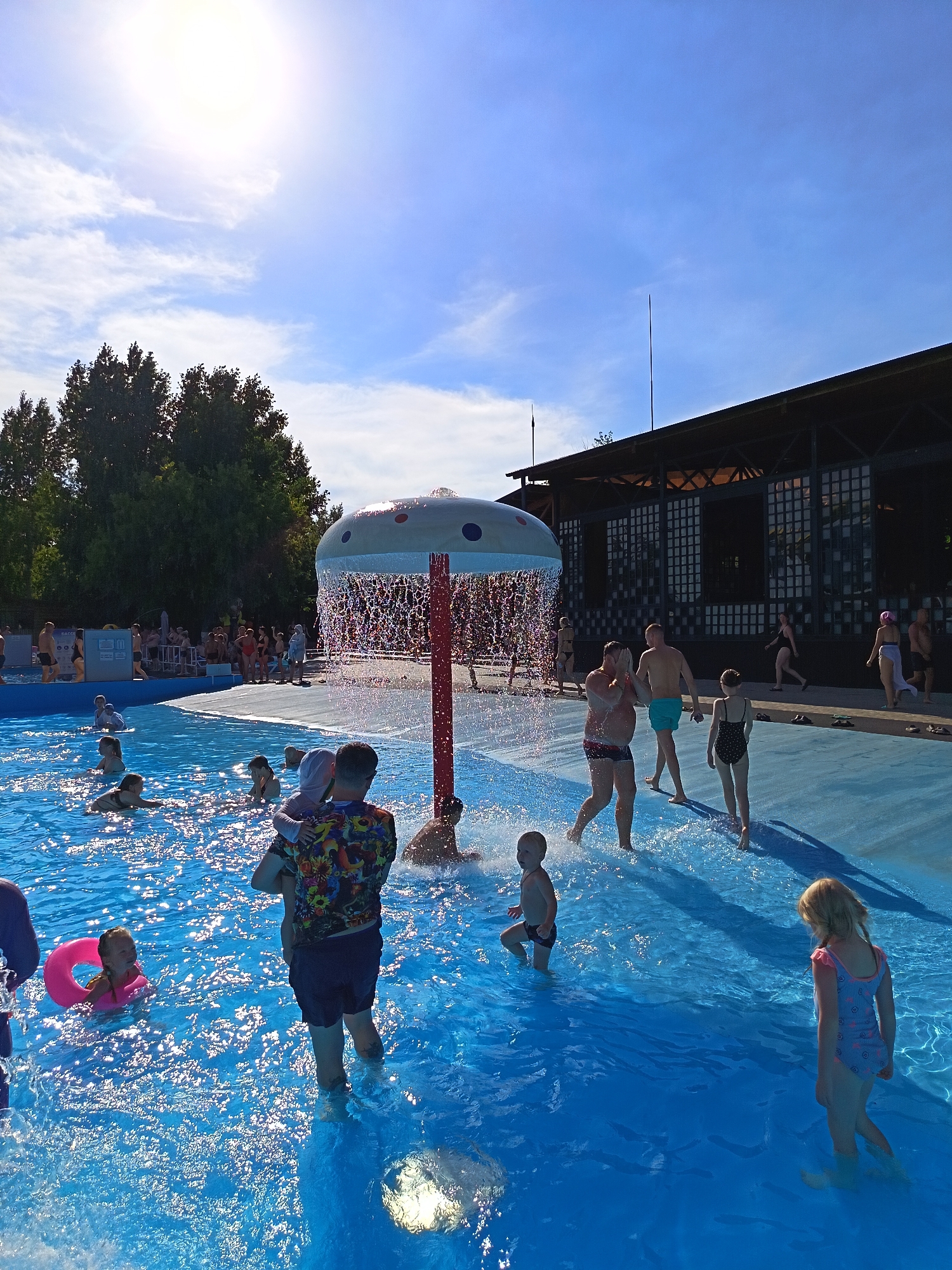 The image depicts a sunny day at an outdoor swimming pool facility in Yarovoye, Russia. In the foreground, the bright blue water of the pool is filled with many people. A young child is being held by an adult in the water. A pink inflatable ring floats near a young girl in the pool. Further into the pool, several other individuals, including children and adults, are visible swimming and playing.
A mushroom-shaped water feature with water spraying down from it is positioned in the middle of the pool. To the right of the water feature, a group of people are standing or walking on a blue tiled area surrounding the pool. Among them is a young girl in a swimsuit and a young boy in swim trunks. In the background, there is a large, dark-colored building with a distinctive architectural style, likely a part of the pool complex. Trees and a bright blue sky with scattered white clouds complete the scene.