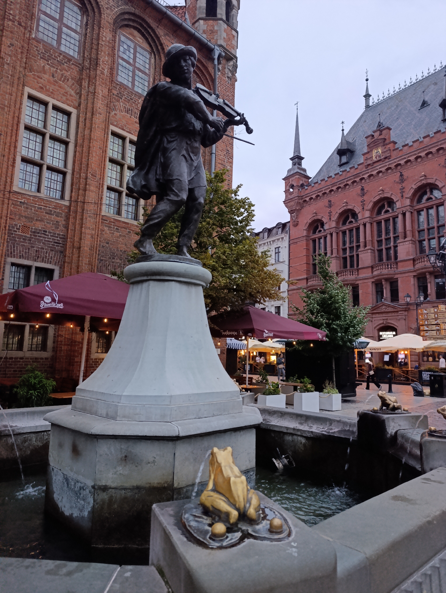 This image captures a street scene in Toruń, Poland. In the foreground, a bronze statue of a person playing the violin stands on a pedestal in a fountain. The fountain features golden frog figurines. The background shows historic brick buildings with arched windows, characteristic of European architecture. Several outdoor seating areas with umbrellas suggest a marketplace or restaurant district. One umbrella displays the text "Pilsner Urquell." Another umbrella has the text "Tinkona" and the logo for "Pilsner Urquell" is visible on another umbrella. The sky is overcast, indicating it might be late afternoon or an early evening with cloudy weather. People are visible in the background, walking through the square and sitting at the outdoor dining areas, suggesting a lively but subdued atmosphere. The overall scene evokes a sense of old-world charm and urban life.