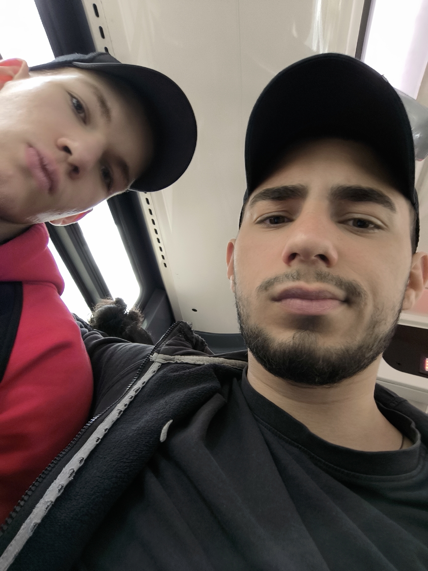 Two young men are posing for a selfie. The man on the right wears a black t-shirt, a black hooded jacket with a white zipper and trim, and a black baseball cap. He has dark hair, a light complexion, and a short, dark beard. The man on the left wears a red hoodie and a black baseball cap. He has fair skin. They appear to be inside a vehicle with a window visible behind them and part of a seat with dark curly hair is visible. The interior has white panels with small, dark ventilation holes. The location, Strasbourg, France is not deducible from the image alone.
