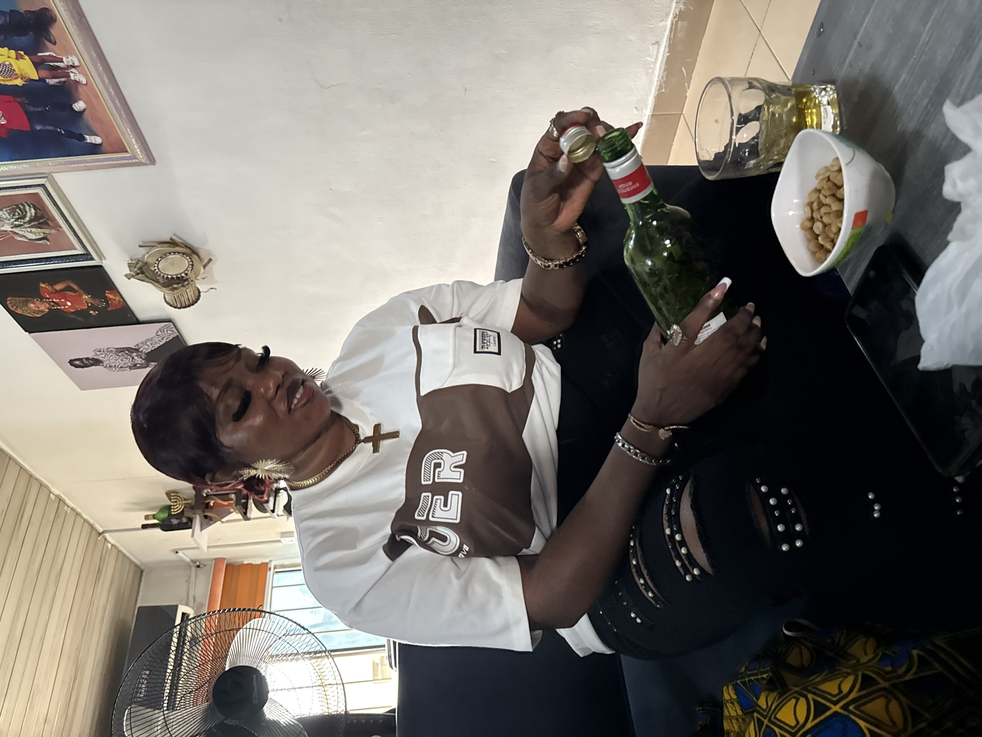 A woman is seated, opening a green bottle of Four Cousins wine at a table in Ado, Nigeria. She wears a white T-shirt with brown accents and ripped black jeans with pearl embellishments. On the table, there is a glass of liquid, a bowl of peanuts, and a smartphone. The room's background includes framed pictures on the wall and a standing fan.
