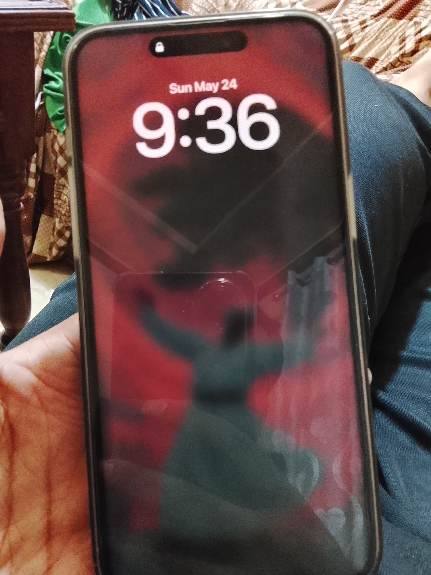 A person is holding an iPhone with a protective case in their hand. The phone's screen displays the time as 9:36 AM on Sunday, May 24th, with a red and black blurred background featuring a silhouetted figure. The person is wearing blue jeans. There is a wooden table leg and patterned cloth partially visible in the background. The location of the image is unknown.