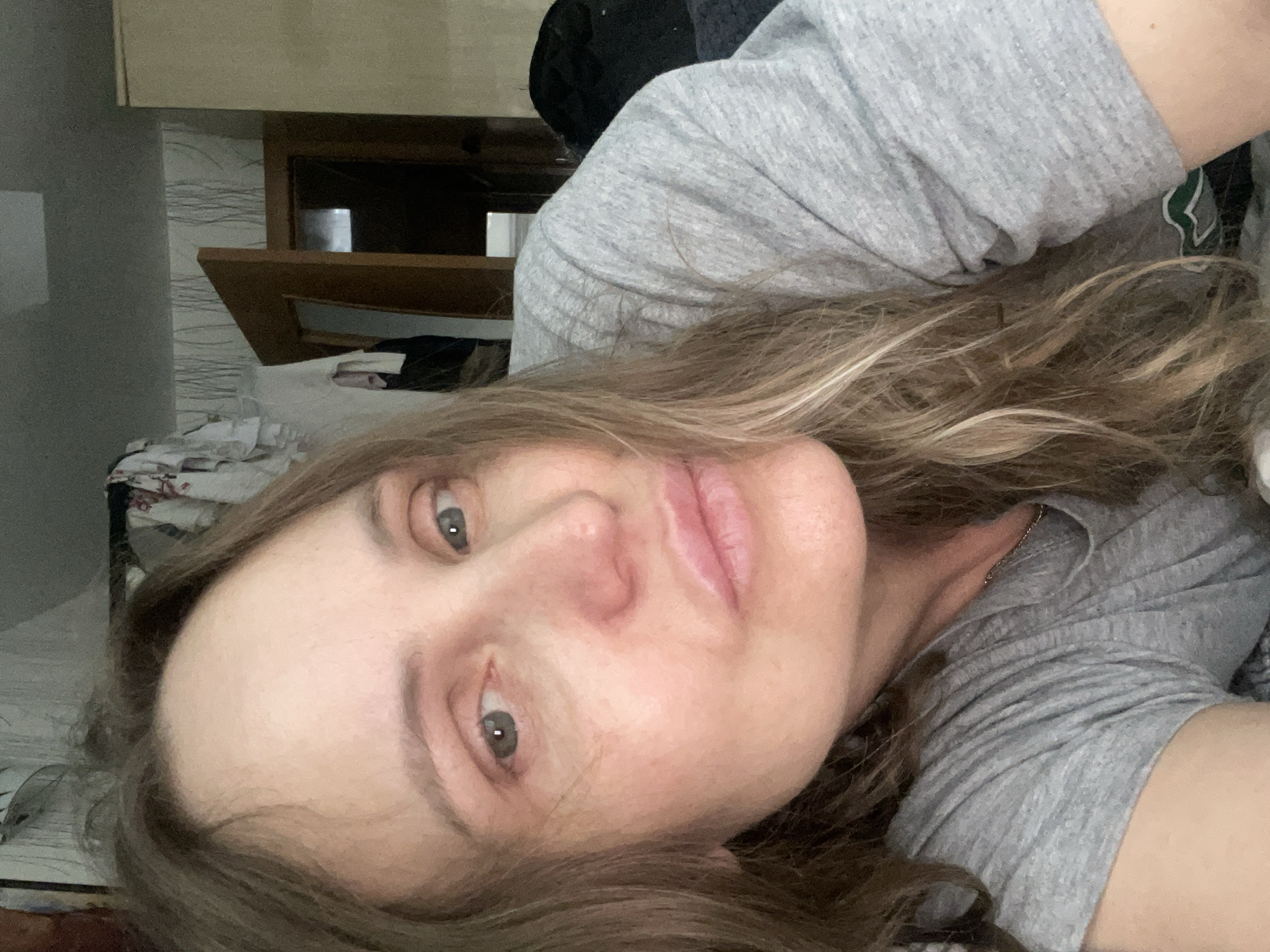 A woman is lying down, facing the camera. She has light brown hair with blonde highlights, fair skin, and light-colored eyes. She is wearing a gray t-shirt. The background includes a piece of furniture with a wooden frame and wallpaper. The location is not discernible to be Chaikovsky, Russia.