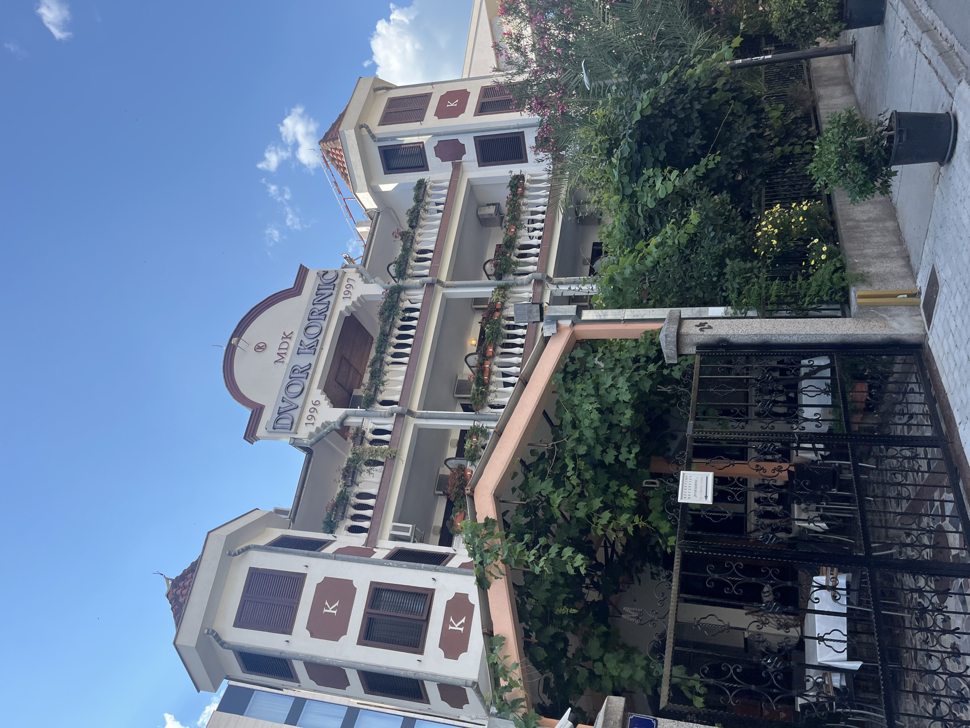 This image shows the facade of a multi-story building with a sign that reads "DVOR KORNIC 1996 MDK 1997". The building has multiple balconies with white railings adorned with potted plants. The architecture features brown window shutters and decorative elements. Lush green foliage, including vines and trees, surrounds the building, partially obscuring some of its details and covering a black wrought-iron fence at the entrance. A sign on the fence indicates "RECEPTION DVOR KORNIC". The sky is clear blue with scattered white clouds, suggesting it is daytime with fair weather. The scene appears to be a street view in Budva, Montenegro, likely a hotel or residential building. There are no people visible in the frame. The overall impression is of a well-maintained property with abundant greenery, set against a bright sky.