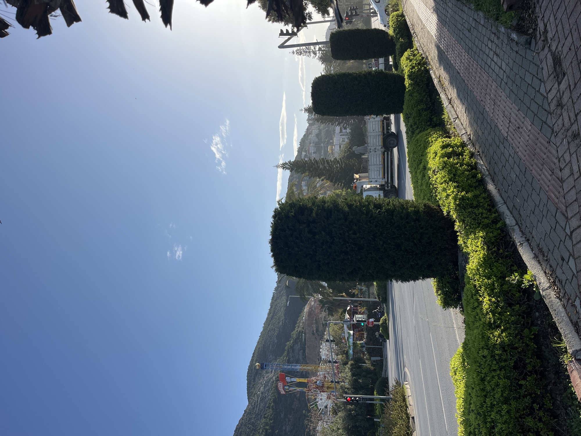 The image is a street-level view in Alanya, Turkey, showing a road running between a sidewalk/paved area and a mountainside. The sky is blue with a few clouds.
The road has a truck stopped along the side. Taller, cylindrical bushes line the road on the sidewalk side. A brick sidewalk runs along the edge.
In the distance, on the mountainside, there's a Ferris wheel and a tall amusement park ride visible. The side of the mountain also shows residential buildings. A traffic light is visible on the left side of the road.