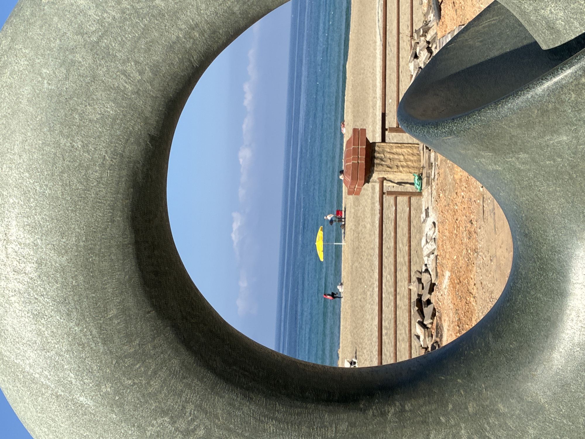 This image presents a detailed view of a beach in Alanya, Turkey, framed through a prominent, dark-grey, textured curvilinear sculpture in the immediate foreground. The sculpture, possibly made of concrete or stone, exhibits a granular surface and forms an arched opening, with a shadowed inner curve.
Beyond the sculptural frame, a light-brown sandy beach extends horizontally. A rustic fence, constructed from wooden posts and horizontal rails, runs across the midground, separating the viewer's immediate area from the main beach. Behind this fence, to the right, a square pillar made of reddish-brown bricks atop a lighter stone or concrete base is visible. The ground directly beneath and behind the fence features reddish-brown earth and a collection of broken grey paving stones, suggesting an area under development or repair. A green object, possibly a hose or tarp, lies near the base of the pillar.
On the beach, several individuals and objects are discernible. A person in a red top and dark bottoms is walking on the sand towards the right side of the frame. In the center-right, a bright yellow beach umbrella stands open. Near the umbrella, a red beach cart or cooler is present, accompanied by two individuals; one is partially obscured, and the other is visible in lighter clothing. Further to the left on the sand, a white beach lounge chair is placed. On the far right, a person is lying down directly on the sand.
The midground transitions into the blue expanse of the Mediterranean Sea, which appears calm, with lighter turquoise hues near the shore and deeper blues further out. The horizon is clearly defined, separating the sea from a clear, bright blue sky dotted with scattered white cumulus clouds, particularly visible in the upper left and central portions of the framed view. Distant, faint darker blue landmasses are visible on the horizon line towards the left. The scene is illuminated by strong sunlight.