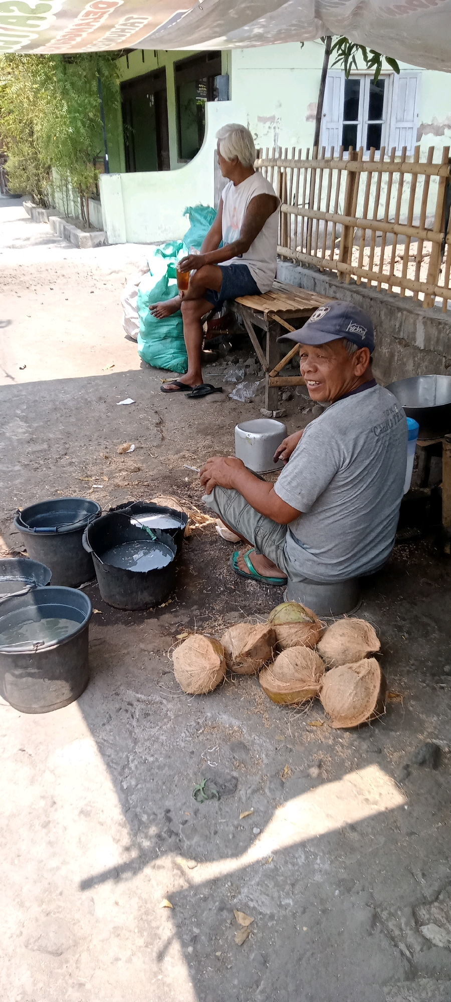 The image depicts a scene outside in Delanggu, Indonesia, likely a street or yard area. Two individuals are present. In the background, an older man with white hair sits on a wooden bench, wearing a white sleeveless shirt and dark shorts. He holds a glass of amber liquid. Next to him are large green sacks. In the foreground, a man wearing a grey t-shirt with text on the back and a blue baseball cap sits on a low grey stool. He is looking towards the camera and smiling. In front of him are several split coconut shells, some whole coconuts, and four dark buckets. Two of the buckets contain a cloudy grey liquid. The ground is paved with concrete and appears dusty with some scattered debris. A bamboo fence is visible behind the seated men. The lighting suggests it is daytime, with shadows cast on the ground.