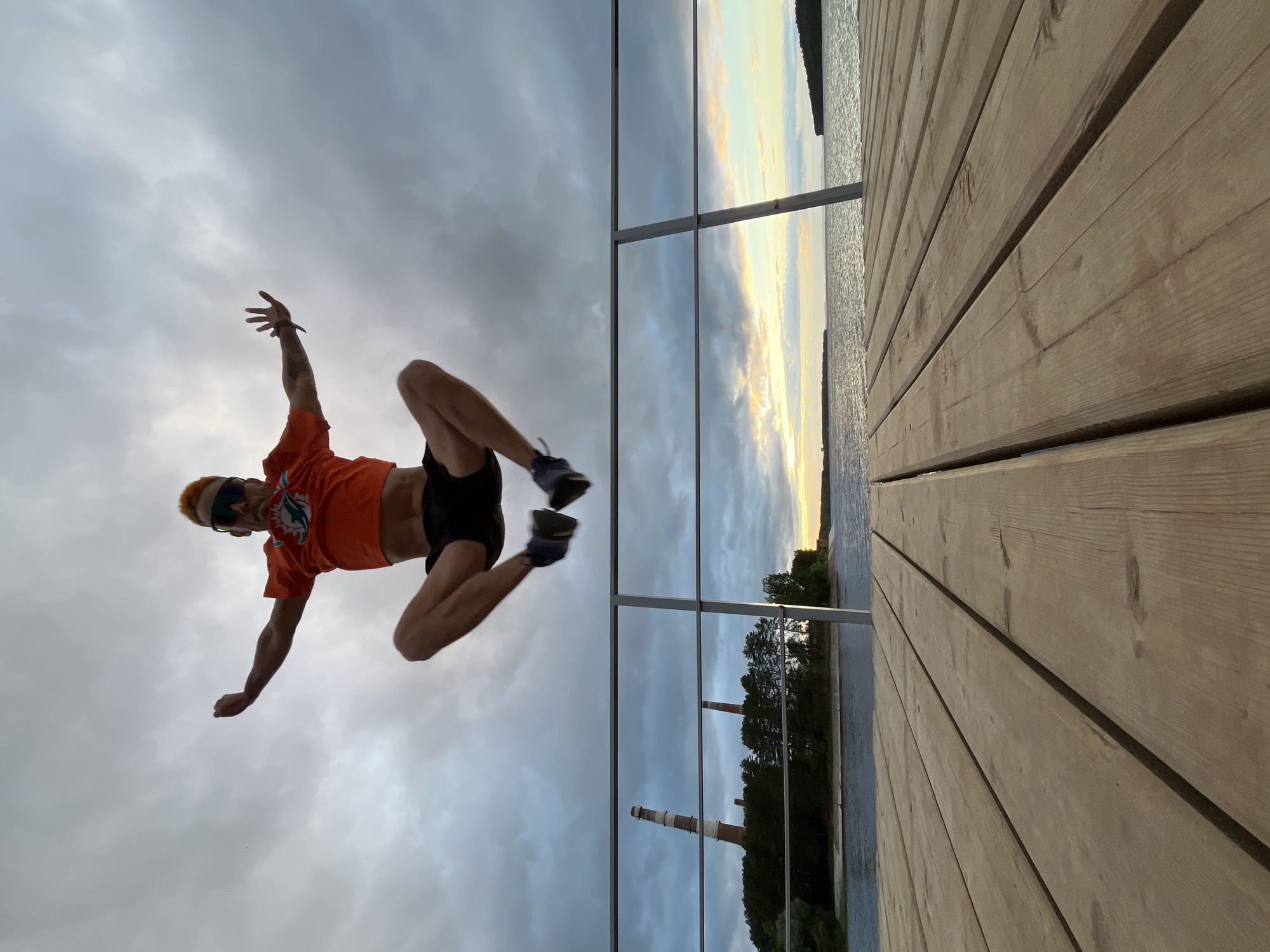 A man with bright orange hair and sunglasses is captured mid-air, jumping with his arms outstretched and legs bent. He is wearing an orange t-shirt with a dolphin logo and black shorts. The photo appears to be a low-angle shot, looking upwards at the man against a cloudy sky.
The image is divided by a reflective surface, possibly a glass railing. On one side, we see the man against the sky. On the other side, the reflection shows the same scene, but also a wooden deck in the foreground, the sky with a sunset glow, a body of water, and a tree-lined shore. Two industrial smokestacks are visible in the distance on the far shore, one red and white and another red.
The time of day appears to be late afternoon or early evening, as suggested by the warm hues in the sky and the long shadows. The weather seems partly cloudy, with dramatic grey clouds contrasting with the lighter parts of the sky. The presence of the wooden deck, the water, and the industrial structures suggest an outdoor setting, possibly a pier or a waterfront area. The man's dynamic pose and the overall composition evoke a sense of freedom and exhilaration.
No visible text is present in the image. The location context provided is Chaikovsky, Russia.