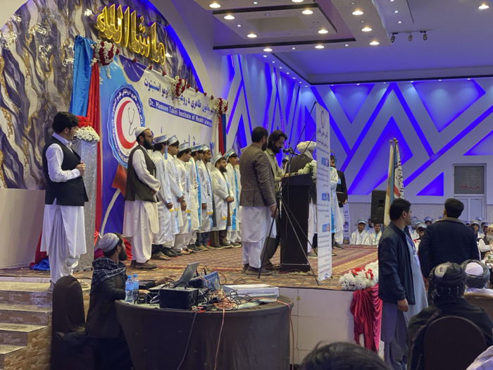 On a stage, a group of men in traditional Afghan clothing and white medical coats stand in a line. Several men at a podium address the audience. In front of the stage is a table with sound equipment. The event appears to be a graduation or ceremony for a medical institute, as indicated by the backdrop with the words "Dr. Mamoon Tahiri Institute of Health Science." Other individuals are seated in the foreground, observing the event. The room has a modern design, with patterned walls and blue lighting accents.