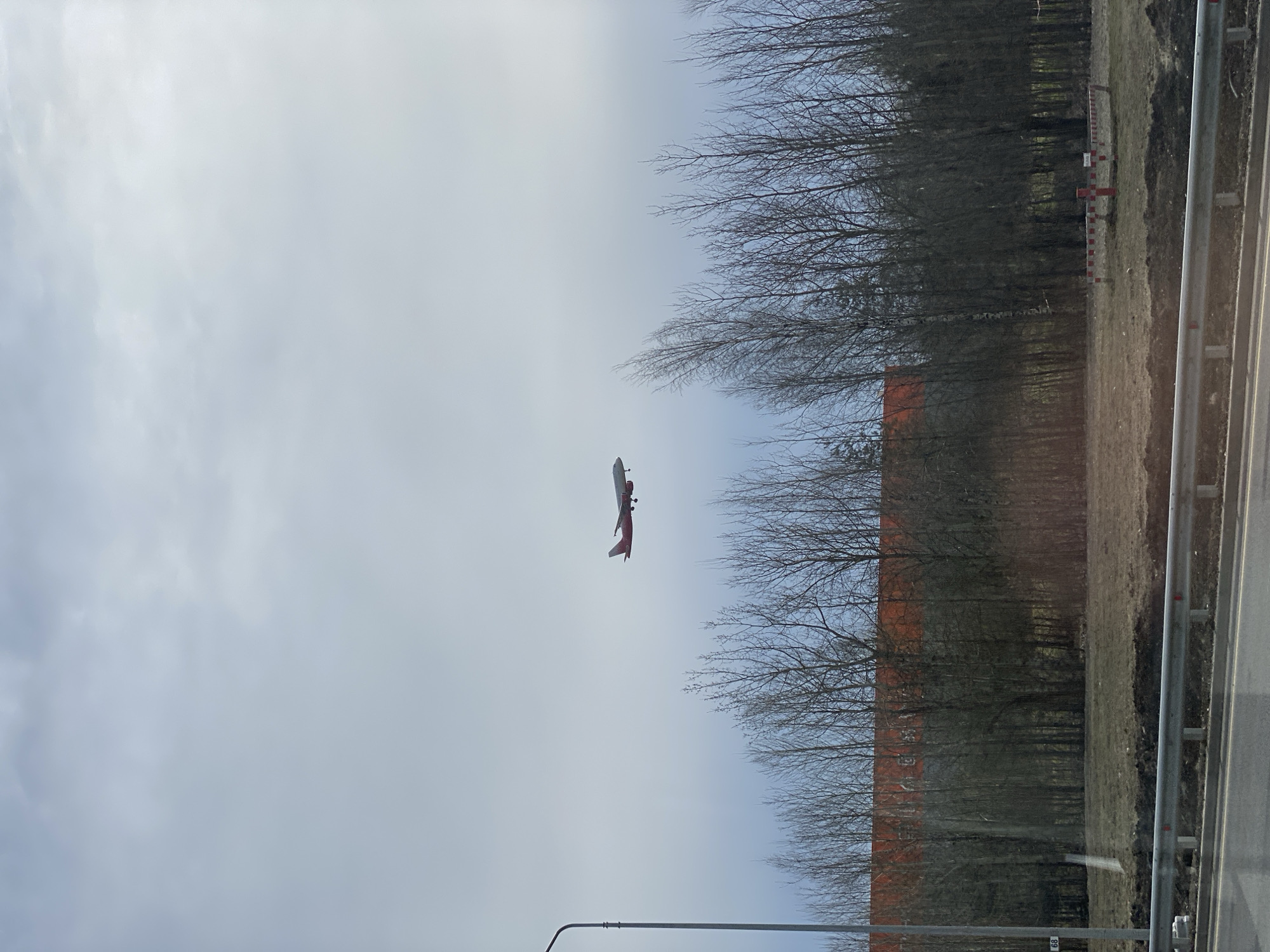 The image depicts a red and white airplane flying in the sky above Saint Petersburg, Russia. In the background, there is a row of trees, a building with a red roof, and a gray sky. A highway guardrail is visible in the foreground.