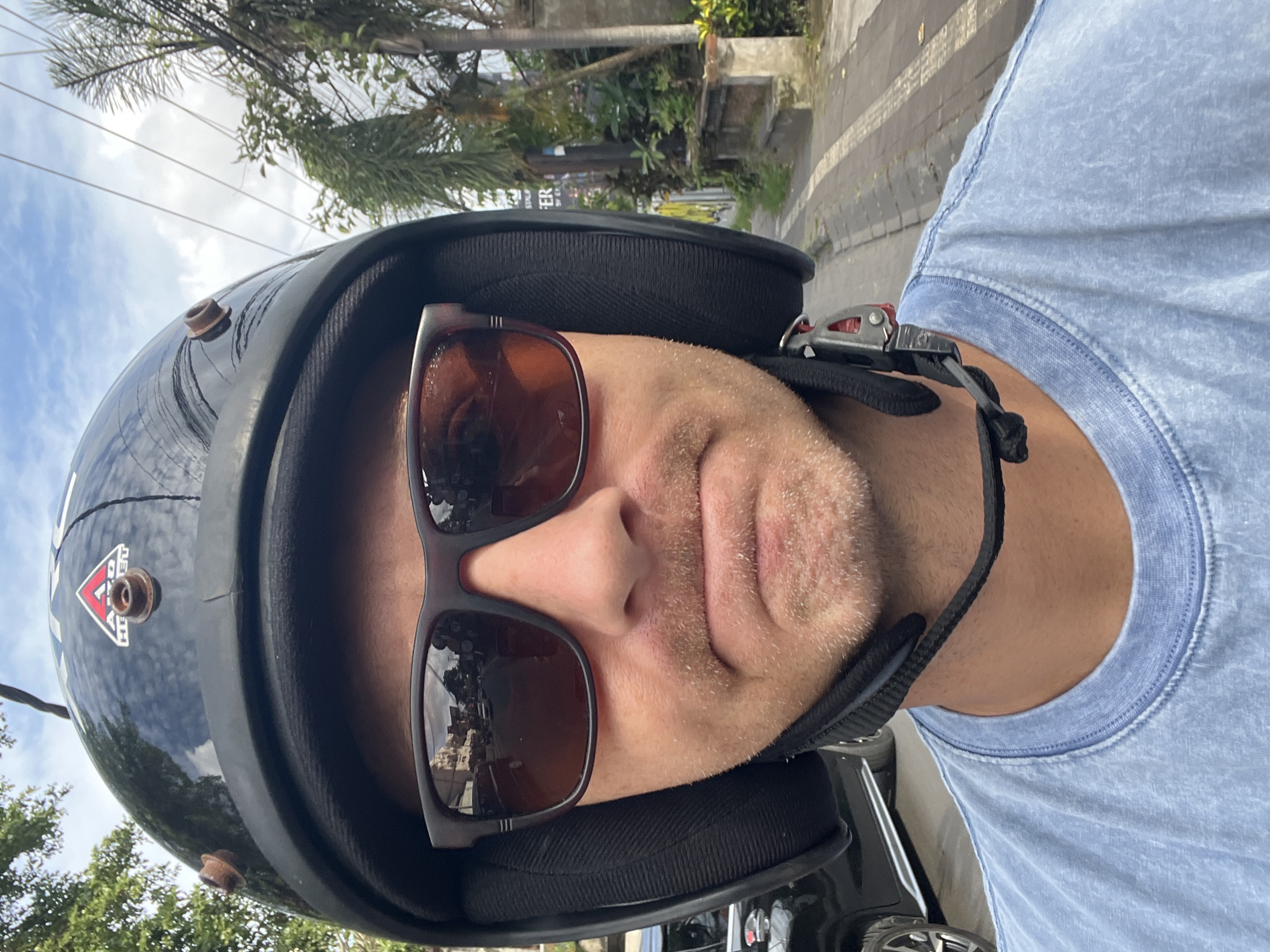 A close-up, upward-tilted shot depicts a person wearing a black motorcycle helmet with a padded interior and chin strap. The helmet features a red and white triangular sticker and protruding metallic fixtures. The person is also wearing brown sunglasses that reflect an urban environment, suggesting the city of Ubud District in Indonesia. The individual has light skin, stubble, and is wearing a blue t-shirt. Background details include trees, power lines, and what appears to be a roadside environment. The person is likely riding a motorcycle or scooter.