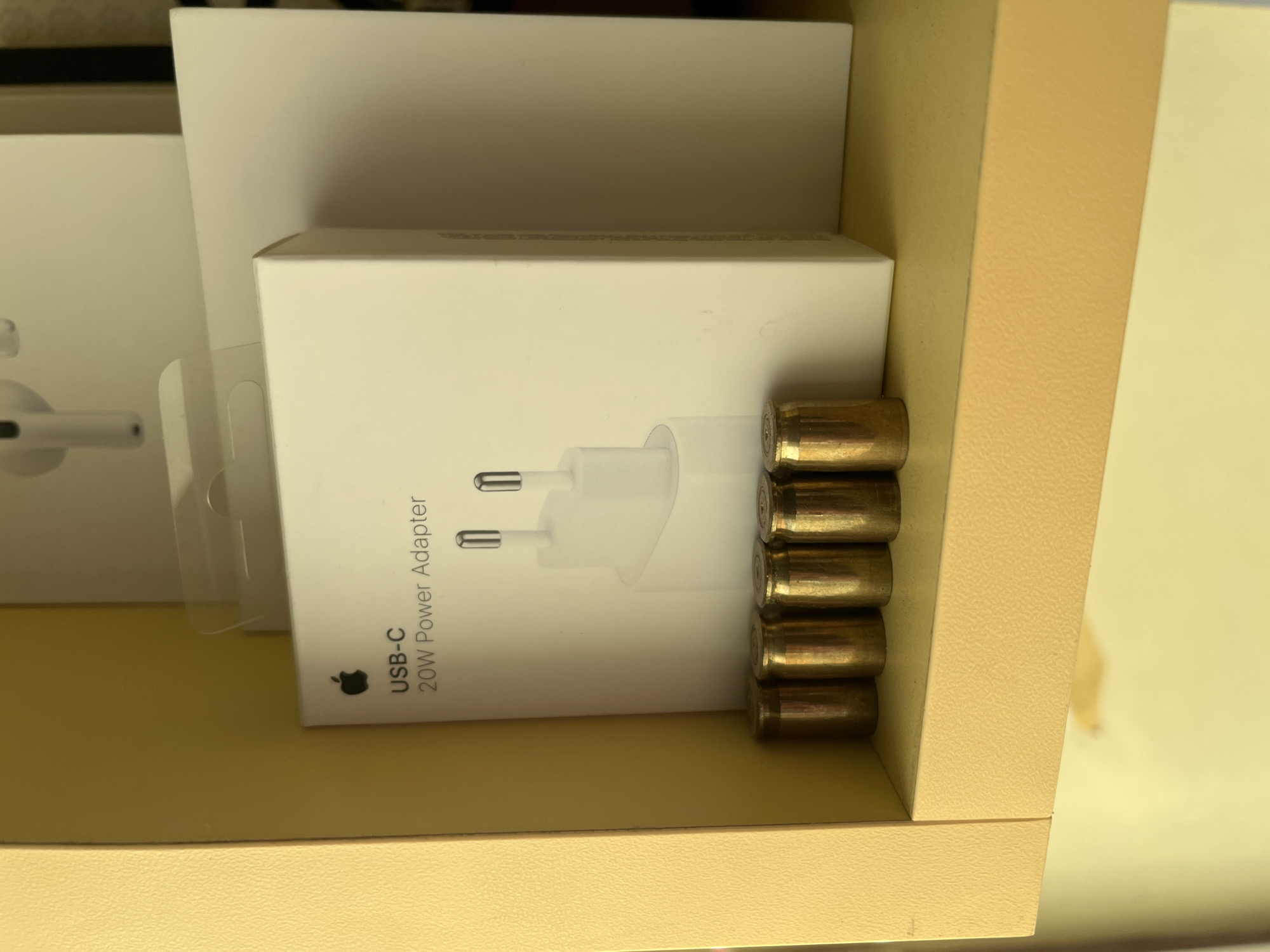 The image, taken in Sofia, Bulgaria, displays a collection of items seemingly stored within a light brown or beige textured drawer or shelf. The dominant subjects are several white boxes, likely packaging for Apple products, arranged vertically.
In the center of the frame, a white box for an Apple "USB-C 20W Power Adapter" is clearly visible, oriented sideways. The Apple logo is discernible next to the text "USB-C 20W Power Adapter". The image on the box shows a white power adapter with two silver prongs, indicating a European plug type. To the left of this box, partially obscured, is another white box, from which a portion of white wireless earphones, resembling Apple AirPods, can be seen. Another white box is visible behind and above the adapter box.
Stacked neatly beside the Apple power adapter box, along the right edge of the frame, are six brass-colored objects. These appear to be either bullet casings or live ammunition rounds, distinguished by their metallic sheen and cylindrical shape. They are stacked horizontally, forming a small column.
The objects are resting on a light beige or pale yellow surface with a subtle texture, which forms the bottom of the storage space. The right side of the image also shows this same textured material, indicating a shelf divider or the side of the drawer. The lighting suggests an indoor setting, and the overall arrangement appears to be a form of organized storage. No human activity is depicted.